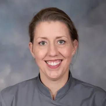 A person with light brown hair pulled back, smiling at the camera while wearing a grey collared shirt