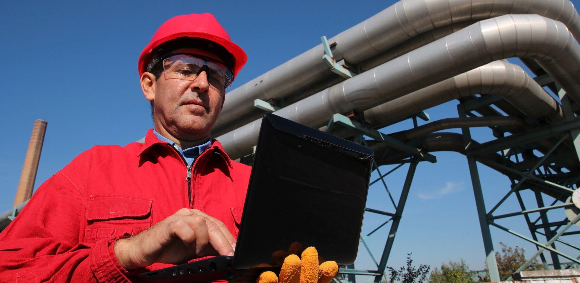 A man in a red hard hat is using a laptop in front of pipes.