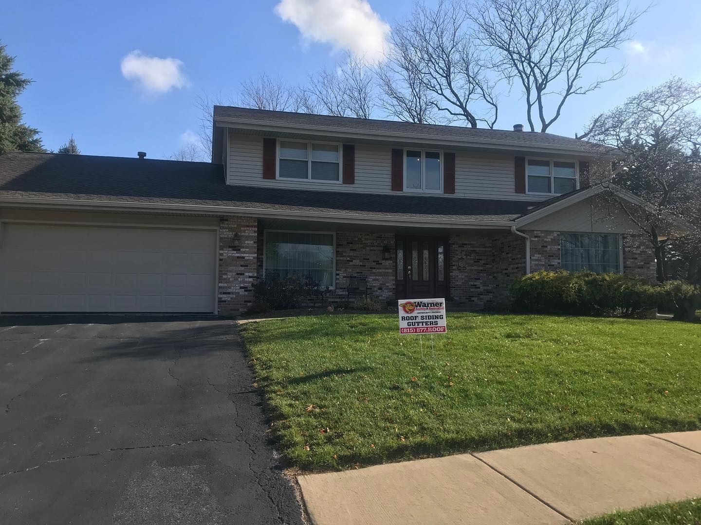 A split-level suburban house with light siding, brick accents, a two-car garage, and a front lawn with a real estate sign.