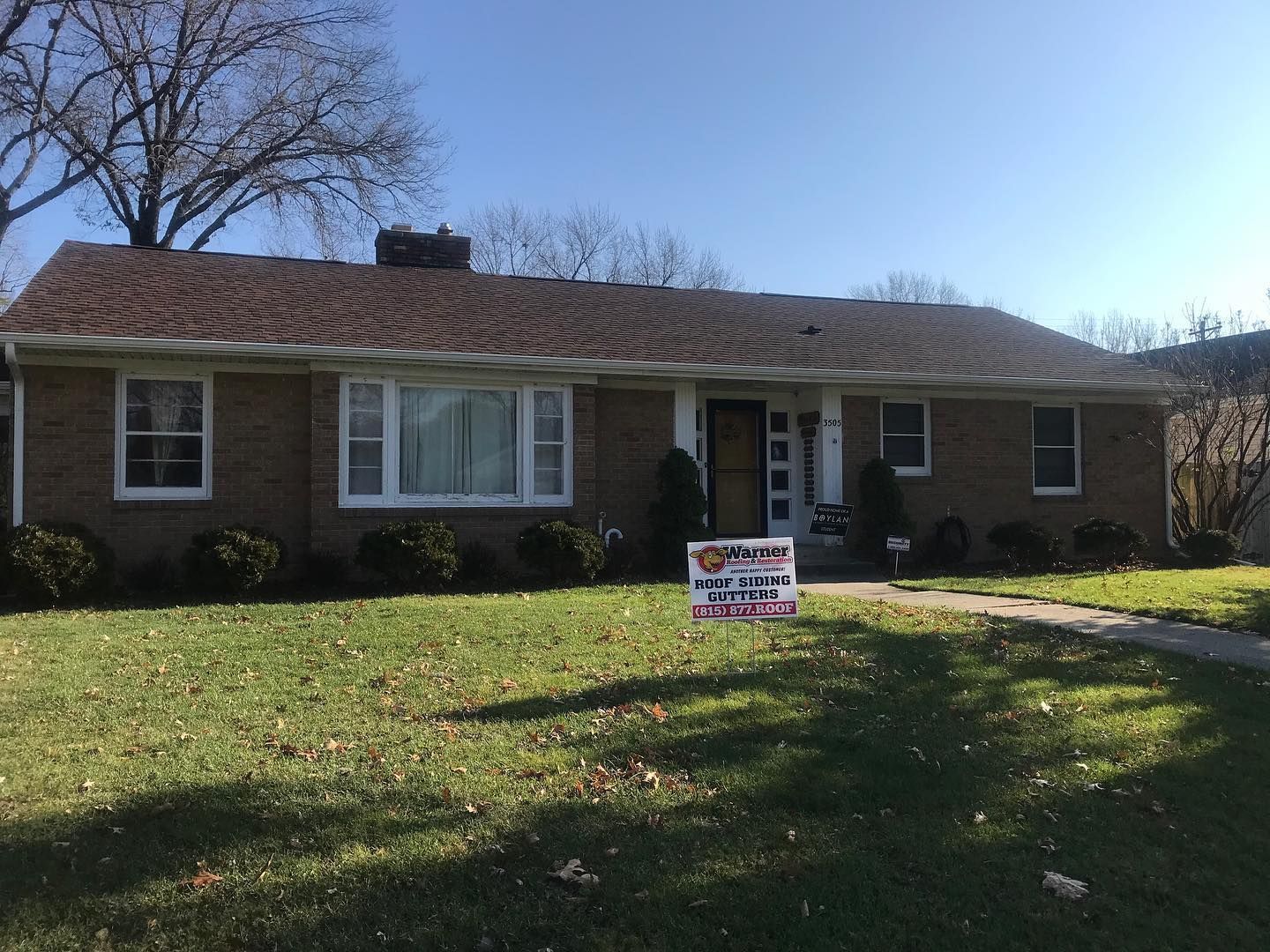 A one-story brick house with a brown shingled roof, lawn, and a small sign in front on a sunny day.