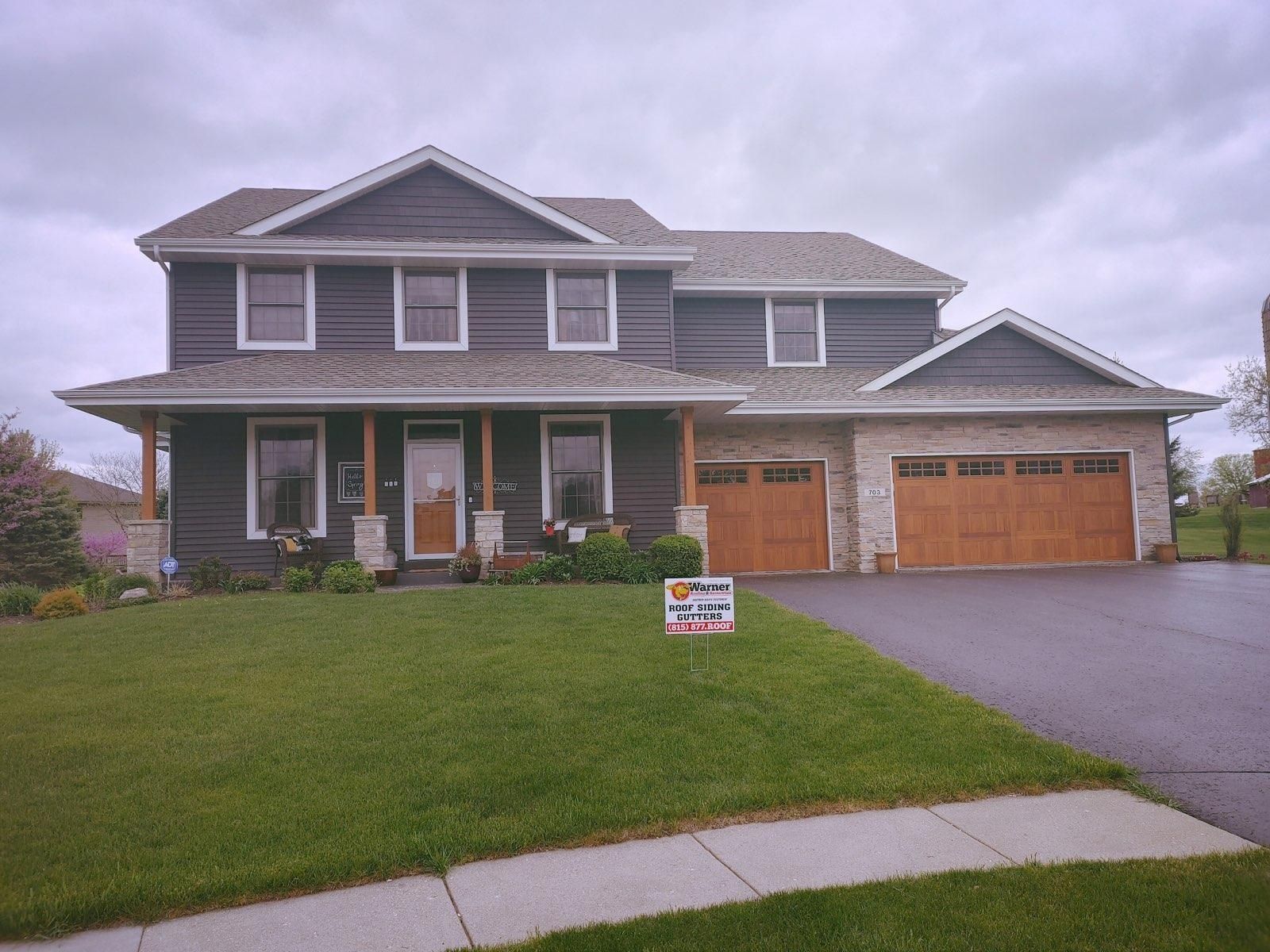 A two-story house with dark siding, a stone facade on the ground level, and two wooden garage doors under a cloudy sky.