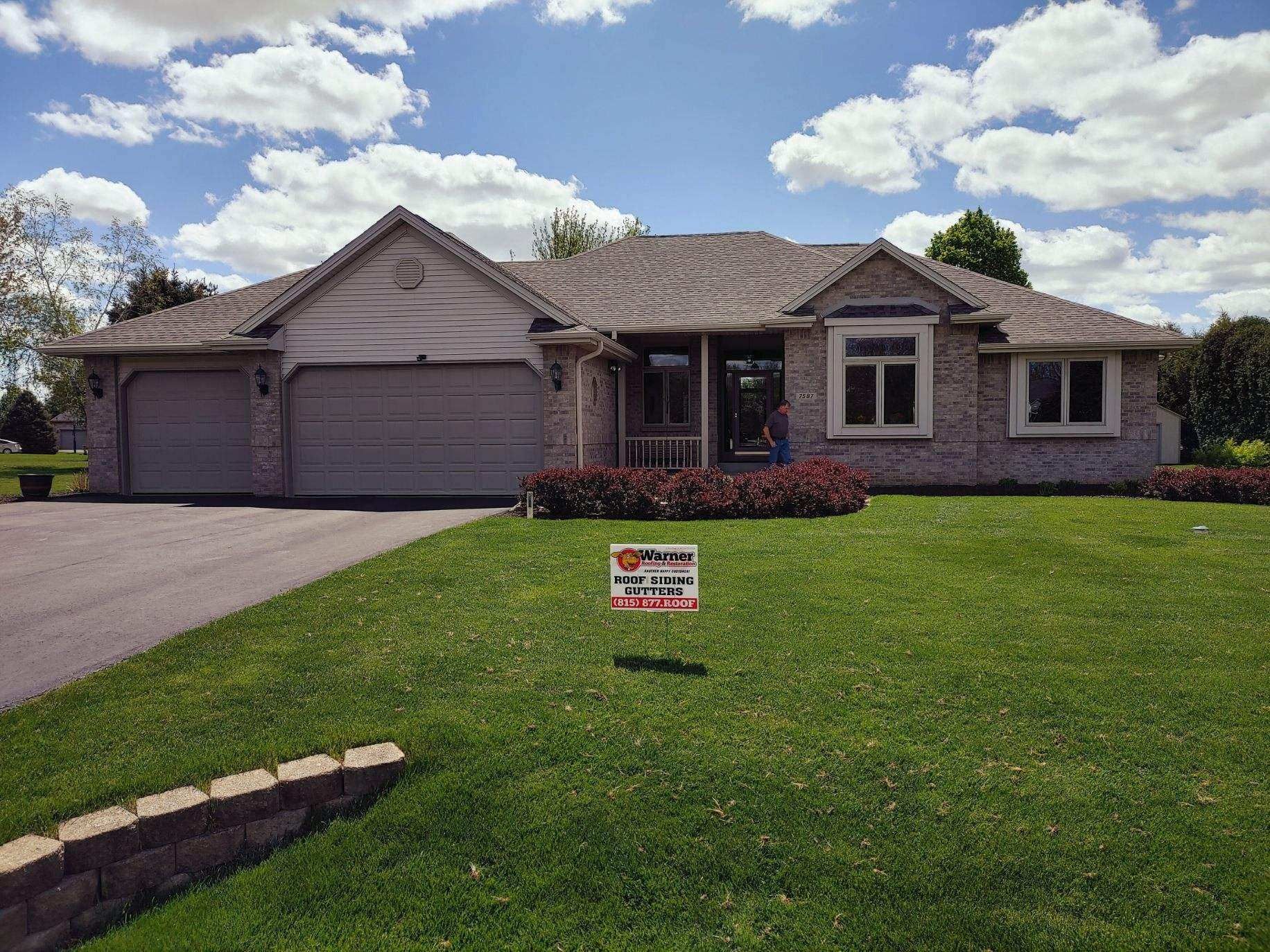 A single-story, tan brick house with a multi-car garage and a paved driveway, seen from a front lawn with a yard sign.