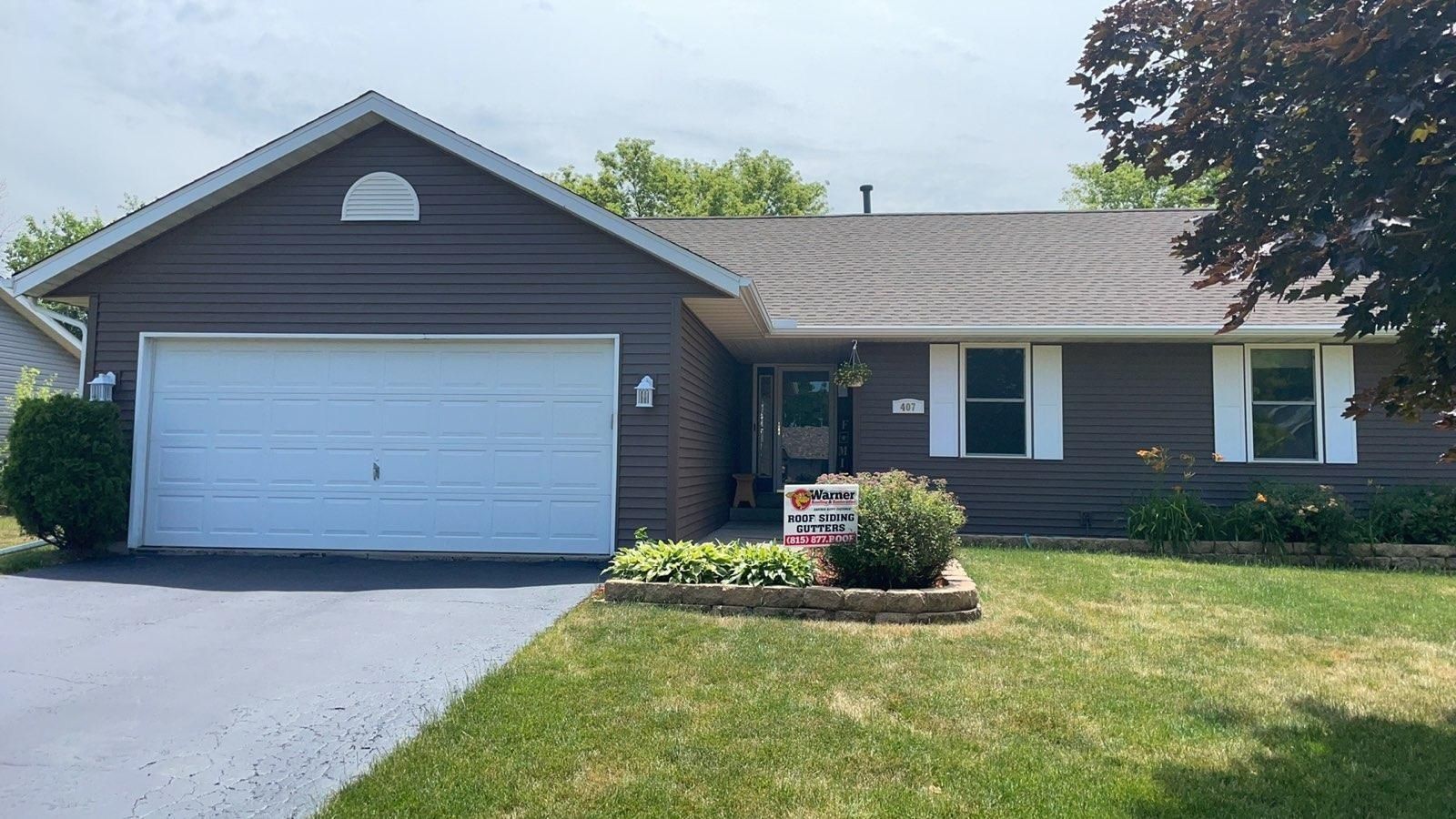 A tan, single-story suburban house with a two-car garage, a small front lawn, and a paved driveway.