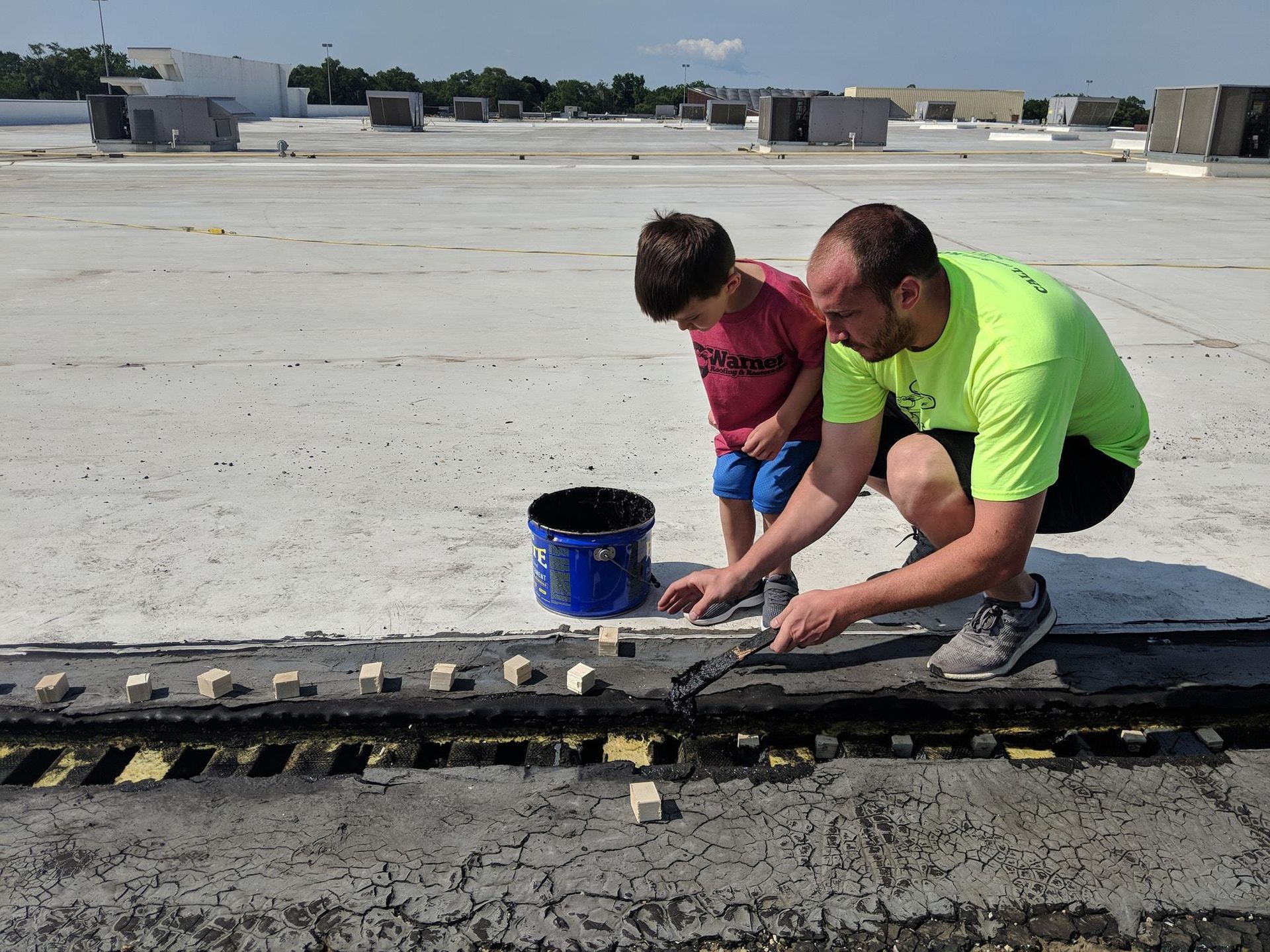 A person in a neon yellow shirt works on a flat white roof with a child nearby, using a tool to place small white blocks.