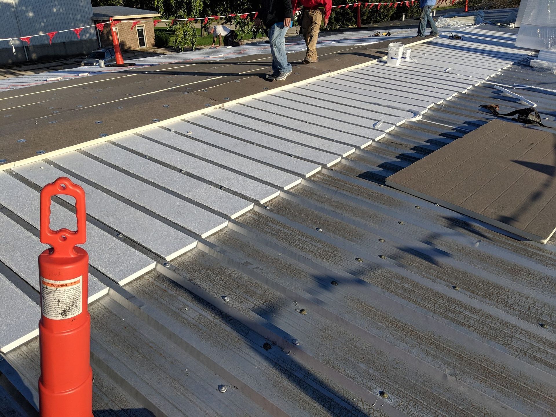 Construction workers install white rigid insulation boards over a metal roof deck, next to a bright orange safety pylon.