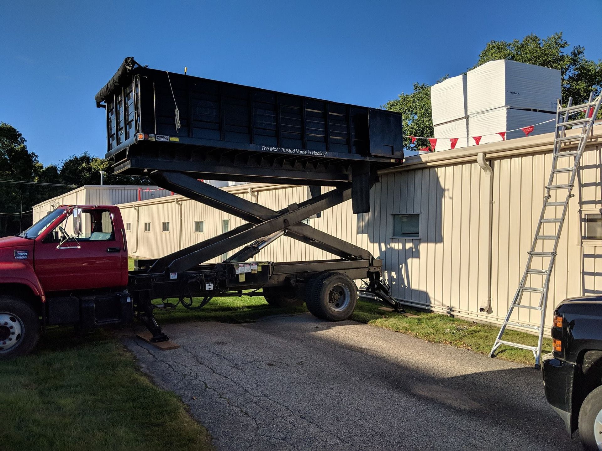 A red truck uses a scissor lift to raise a black dump trailer to the roof level of a light-colored commercial building.