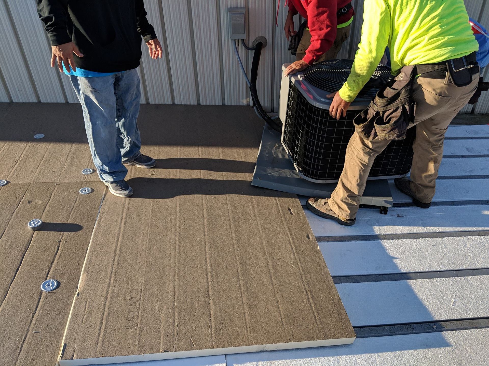 Two workers install a cooling unit on a flat roof next to a panel on a textured surface.