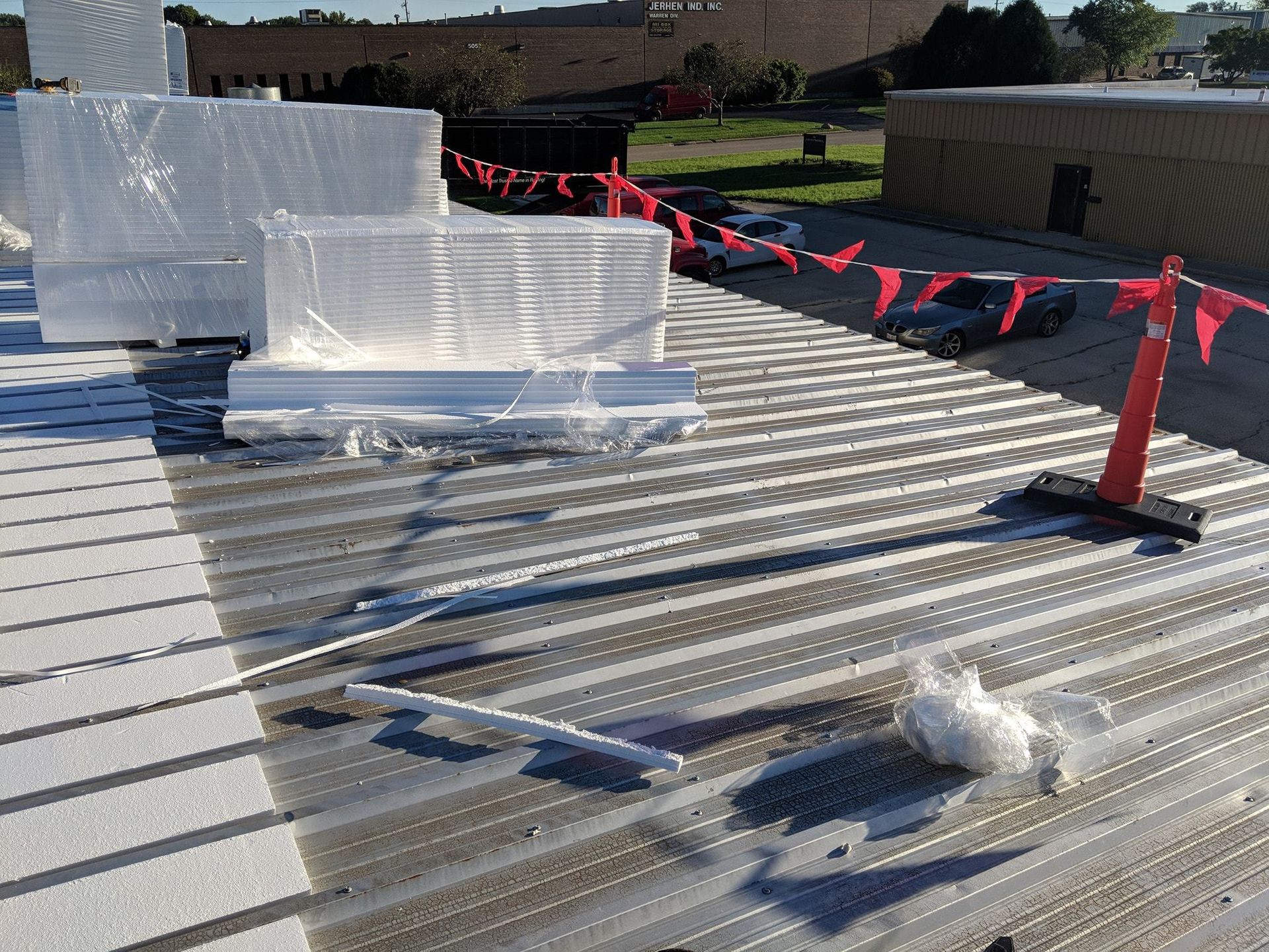 Stacks of construction insulation wrapped in plastic on a corrugated metal roof near a red safety cone and warning flags.