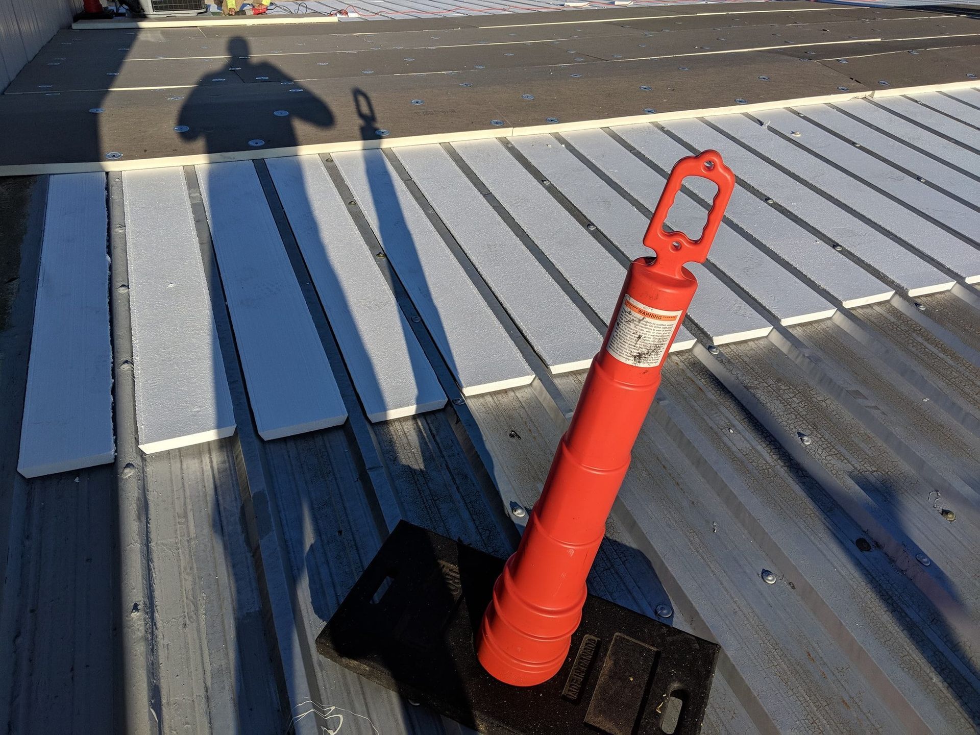 A retractable orange safety cone on a black base sits on a corrugated metal roof with white foam insulation panels.