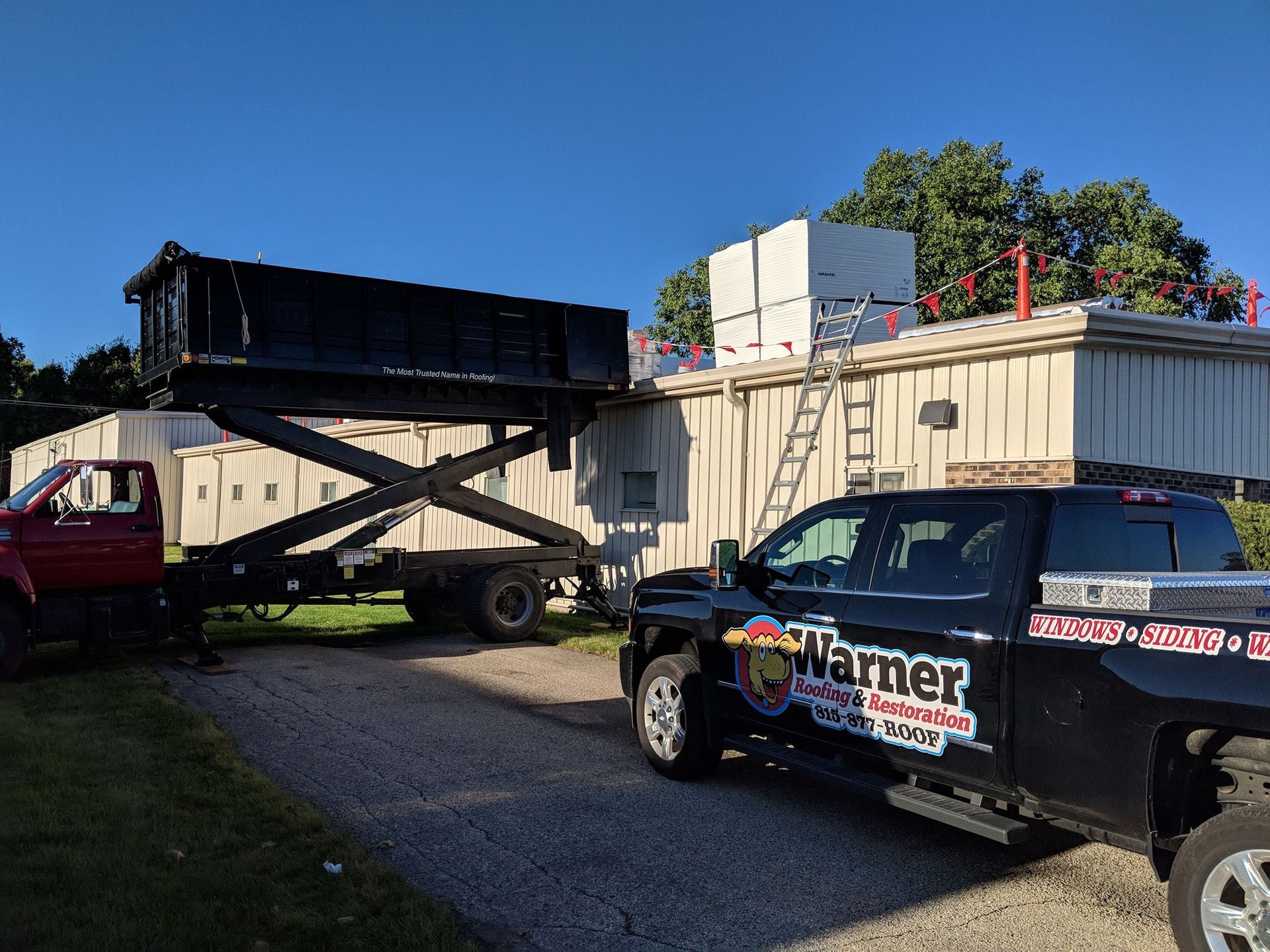 A black dump trailer raised on a scissor lift next to a building with roofing materials and a work truck parked nearby.