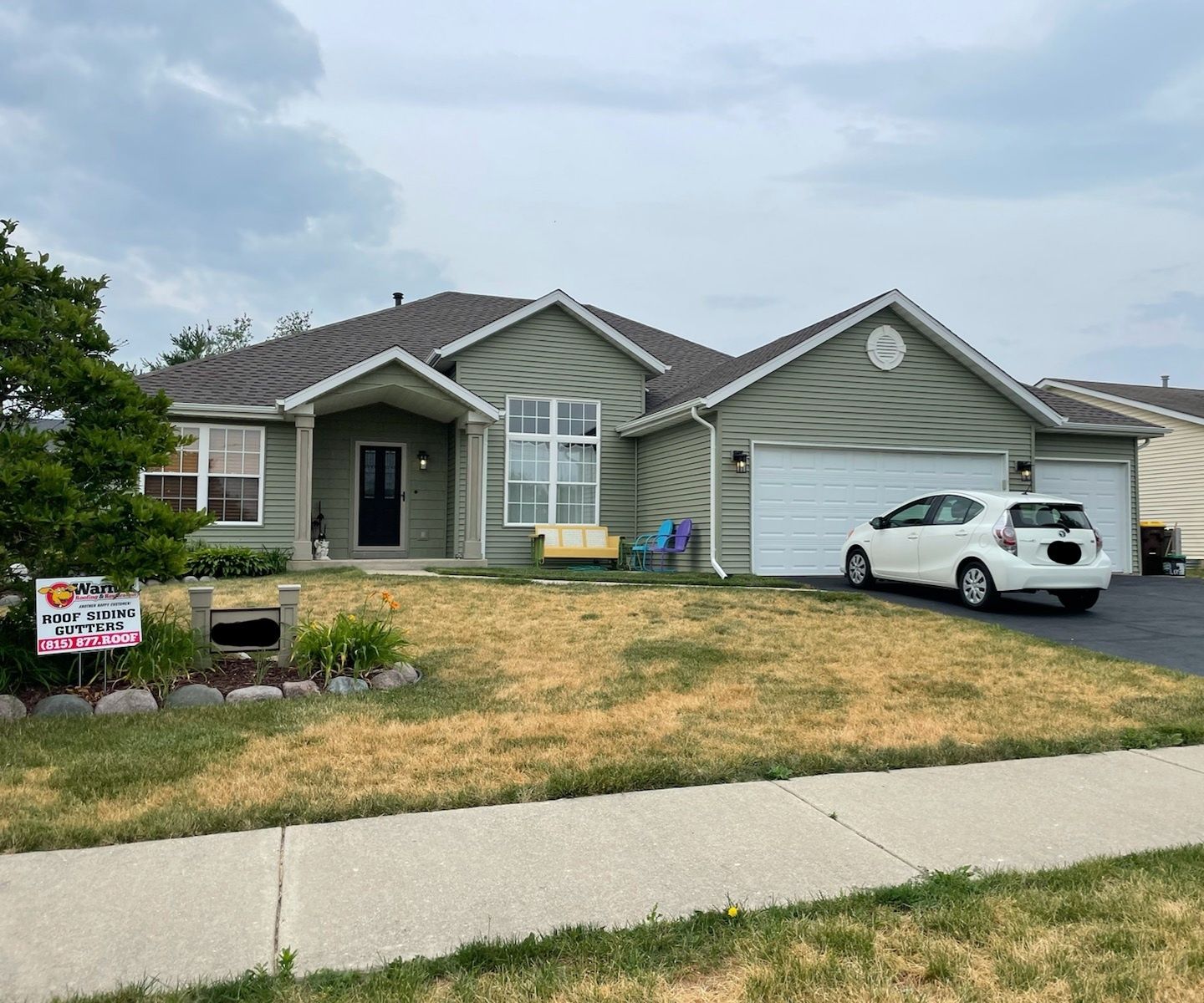 A single-story, sage green suburban house with a white garage, a dark front door, and a white car parked in the driveway.
