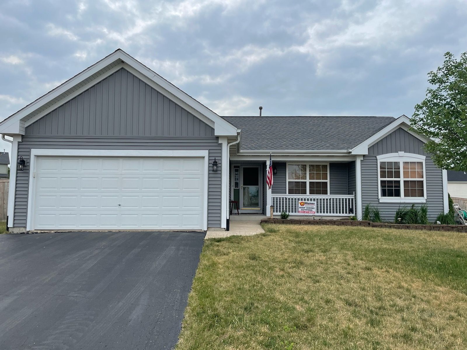A one-story gray house with a two-car garage, white trim, a front porch, and a paved driveway under a cloudy sky.