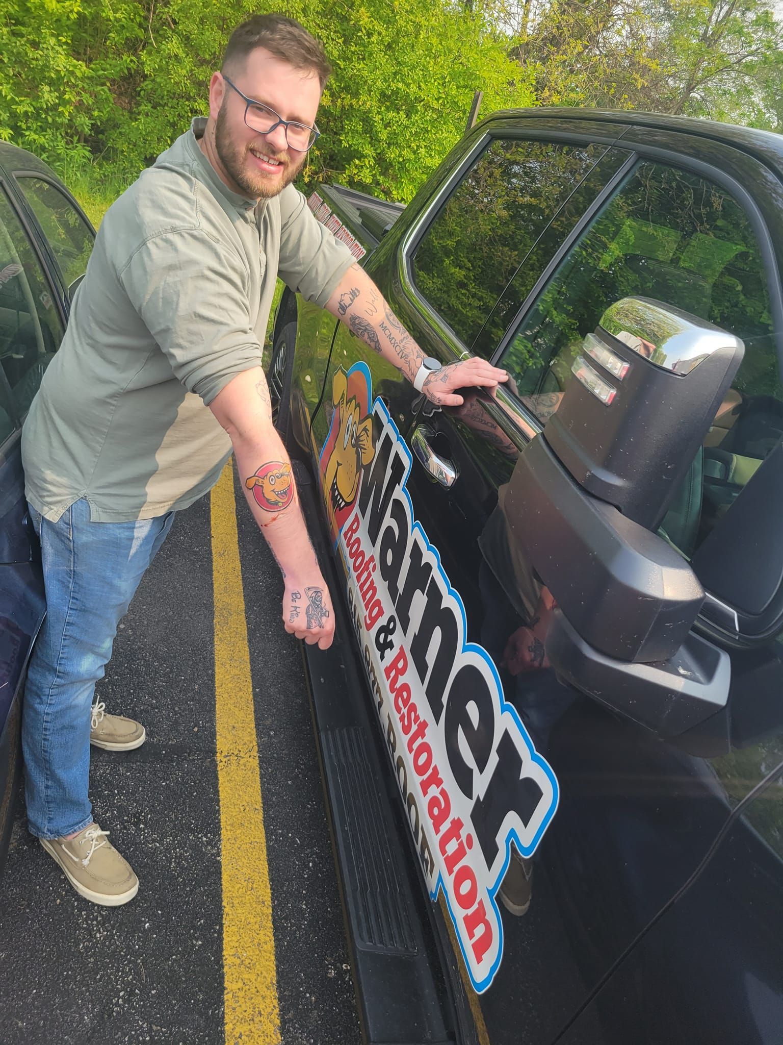 A smiling person in a green shirt and jeans leaning against a black work truck with 