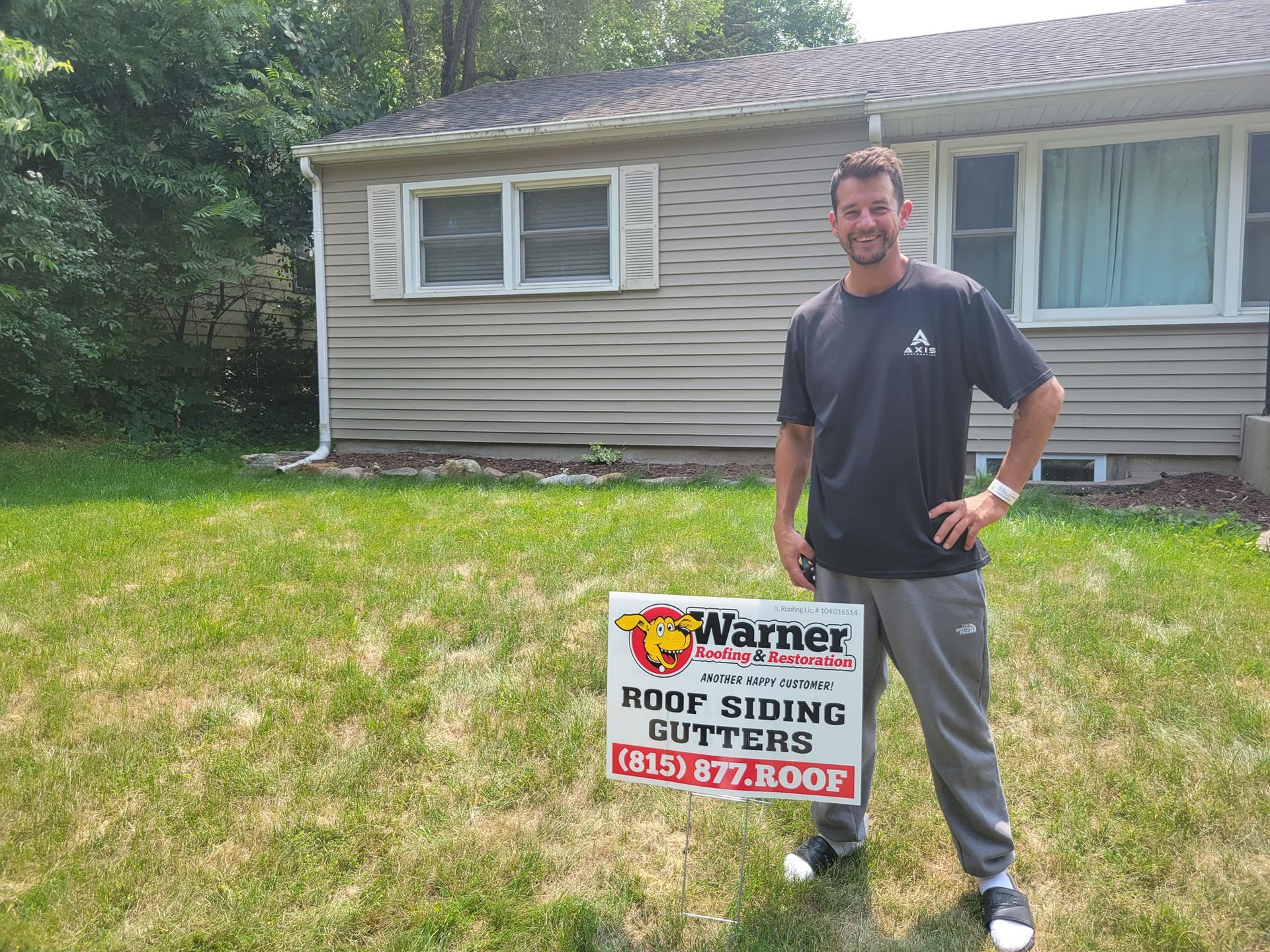 A smiling person holds a Warner Roofing & Construction sign in front of a house with tan siding and a white window.