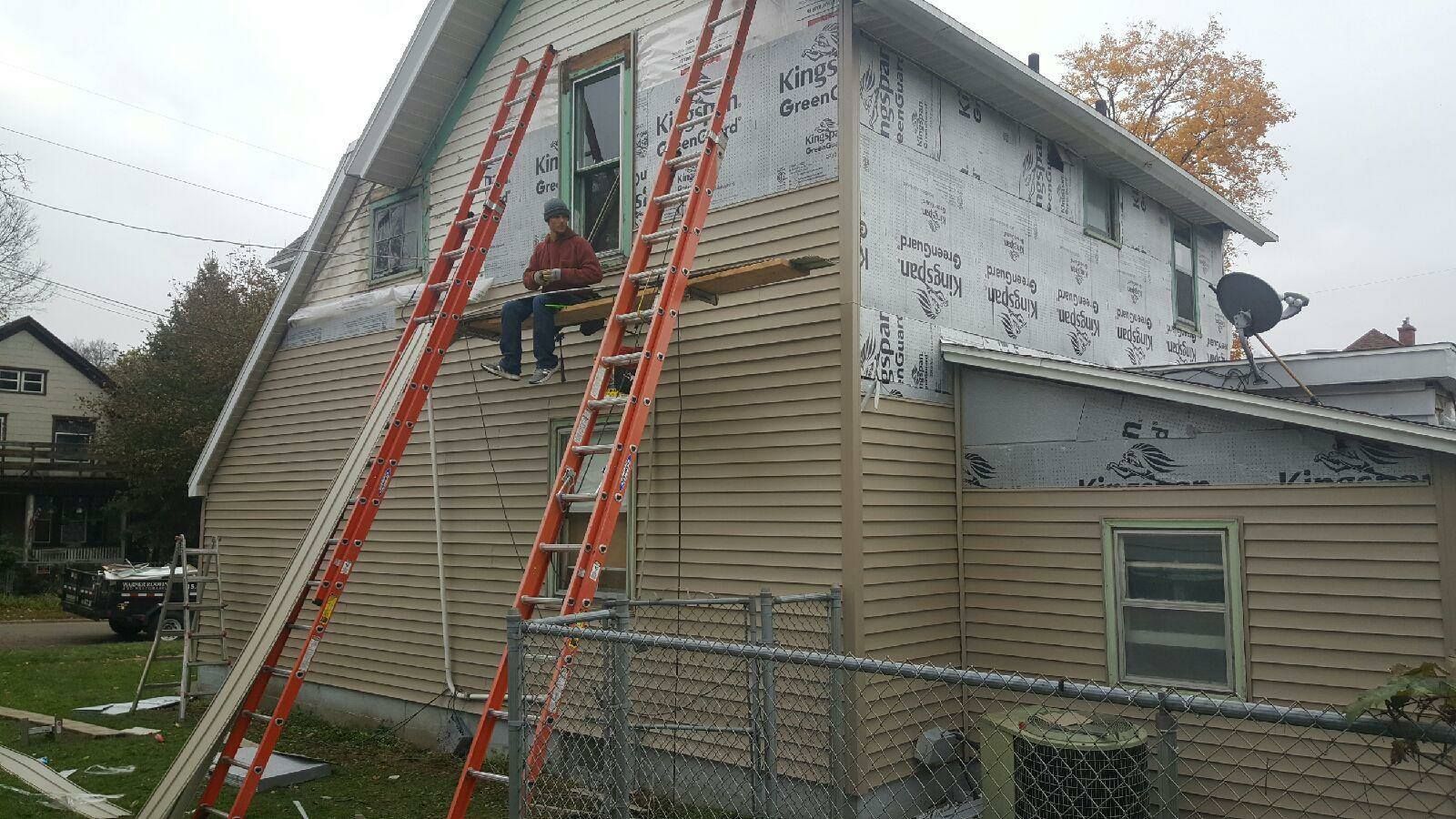 A person sits on a wooden plank supported by two red ladders against the side of a house undergoing siding installation.