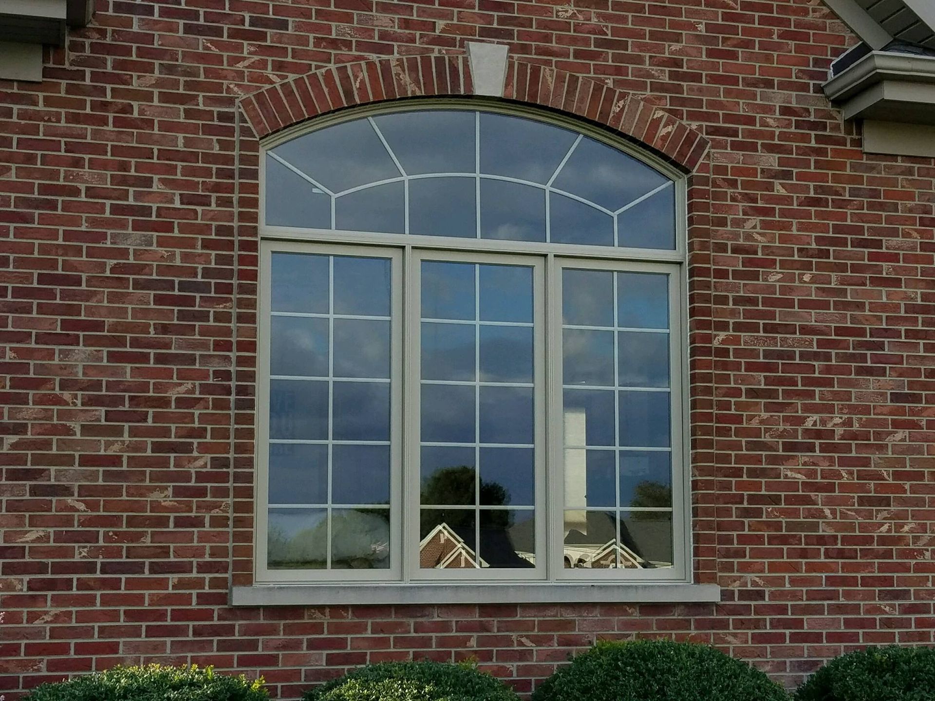 A three-pane arched window with a white grid frame set into a reddish-brown brick wall.