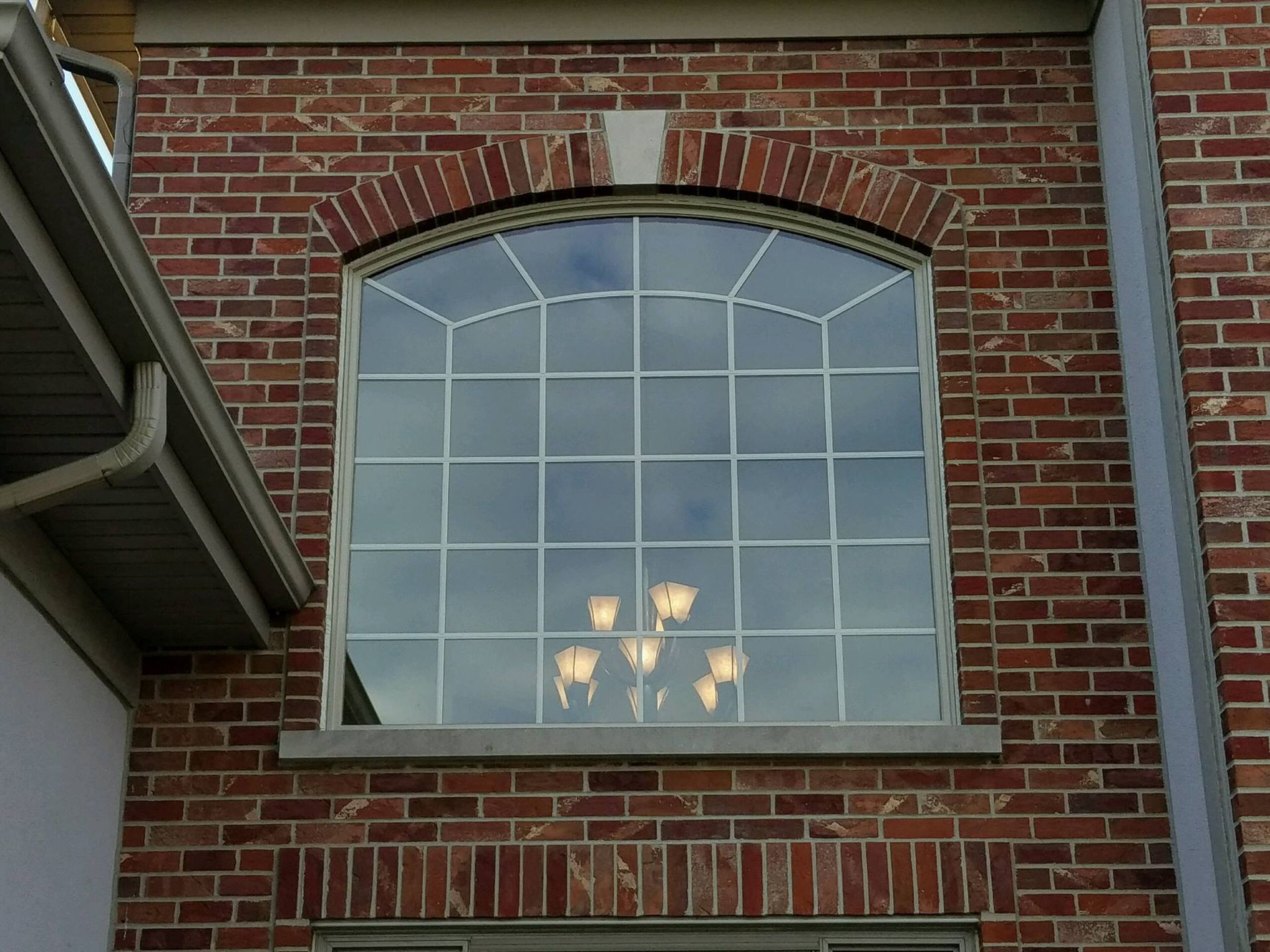 A large arched window with a grid pattern on a red brick exterior wall, showing a chandelier reflected in the glass.