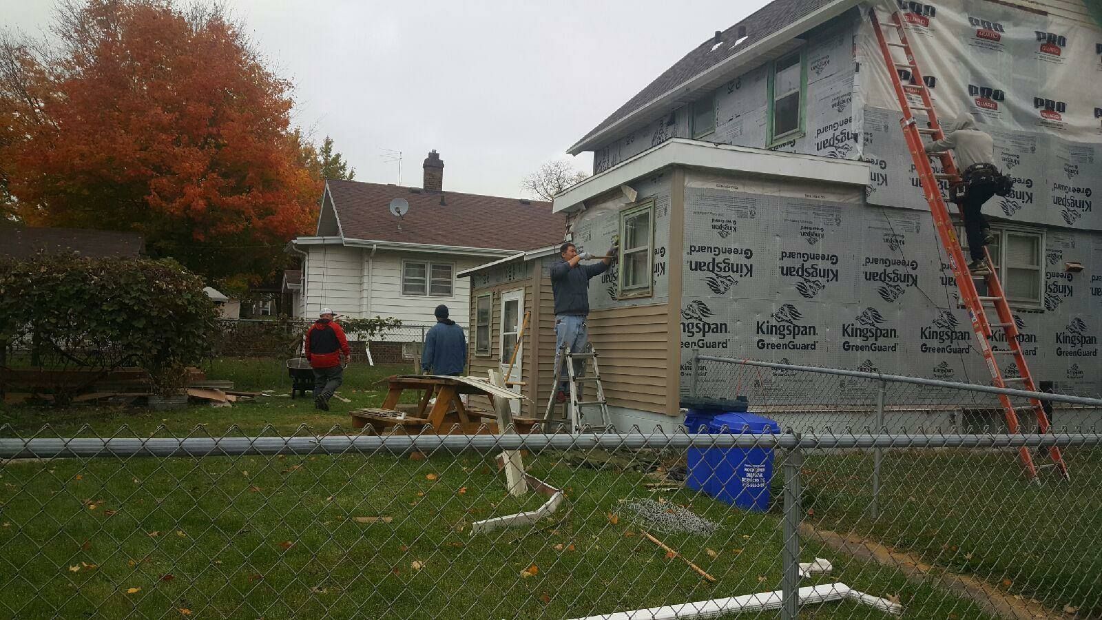 Three construction workers repair the exterior of a house, using a ladder and scaffolding on a suburban lawn.