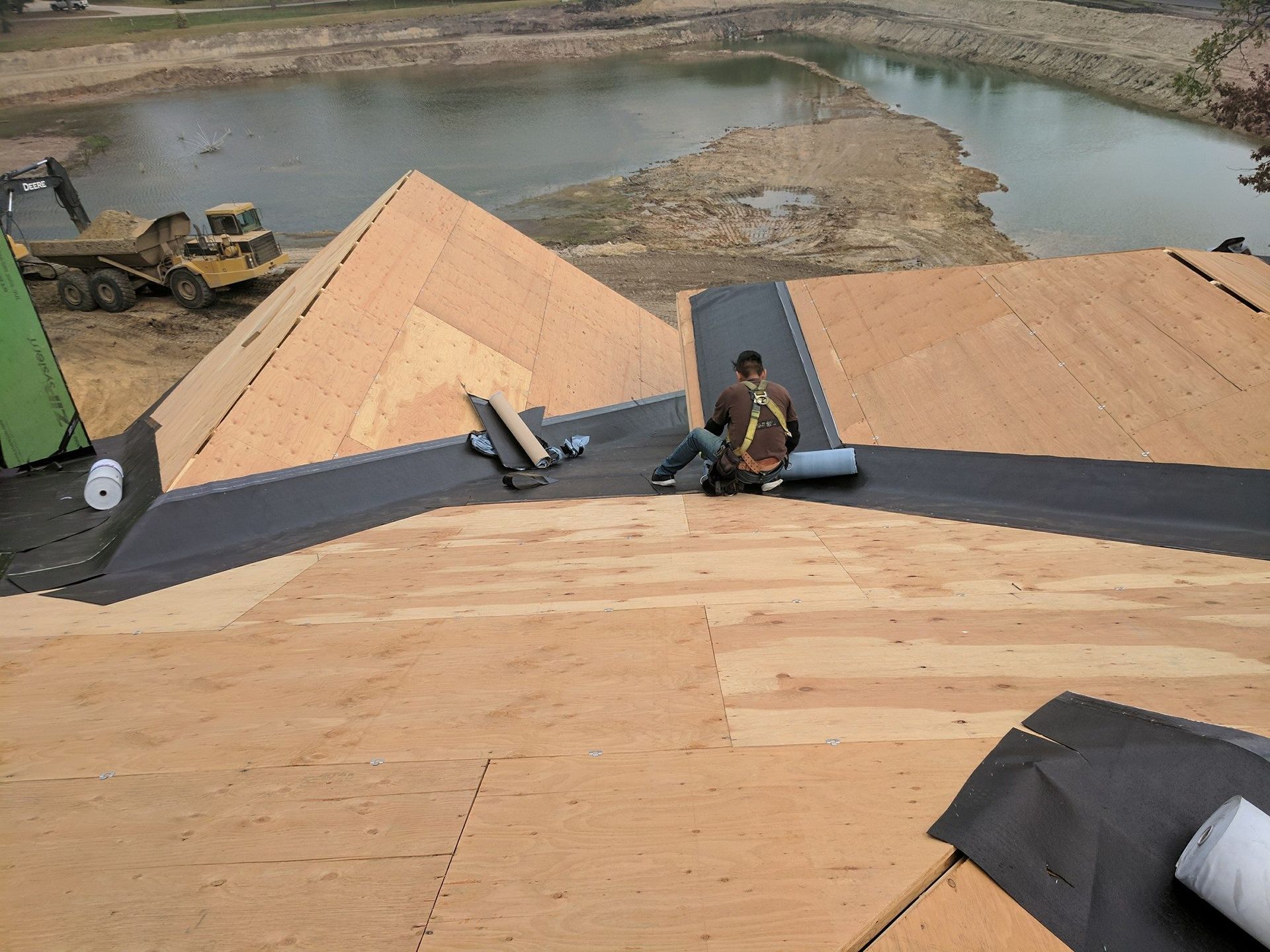 A construction worker wearing a safety harness installs black underlayment on a wooden roof overlooking a pond.