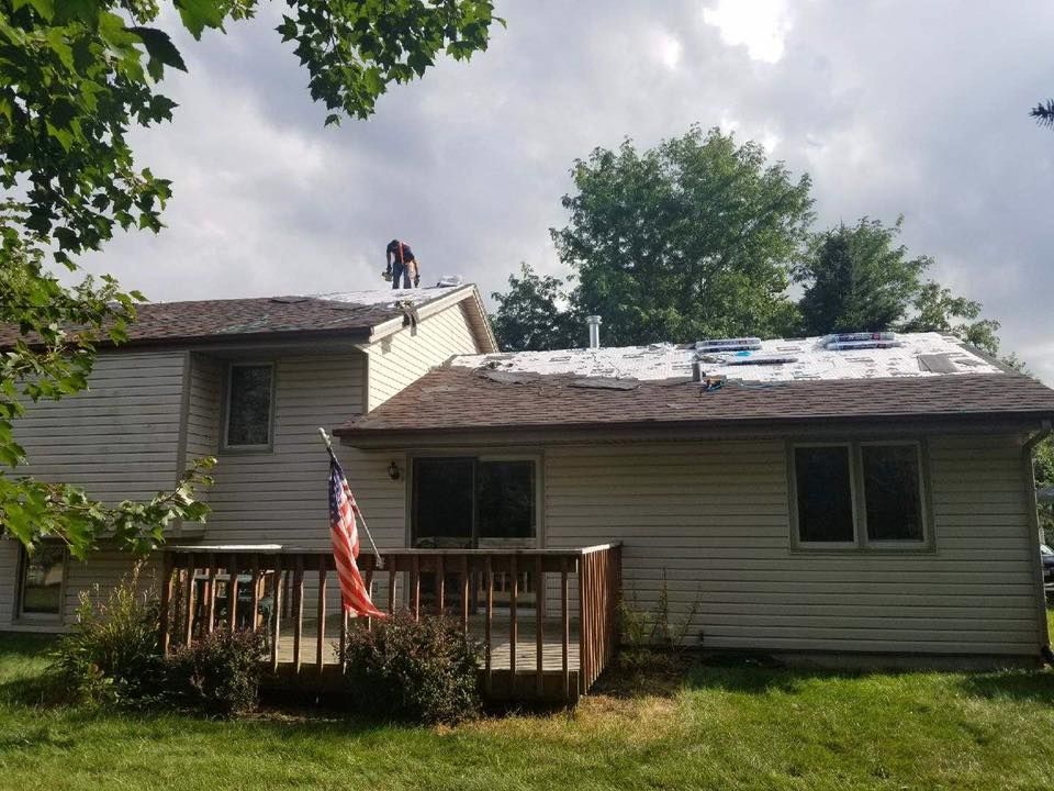 A person works on the roof of a two-story house with beige siding, a wooden deck, and an American flag in the backyard.