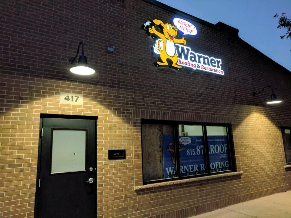 Brick storefront for Warner Roofing & Restoration with a lit sign featuring a dog mascot and address 417 above the door.