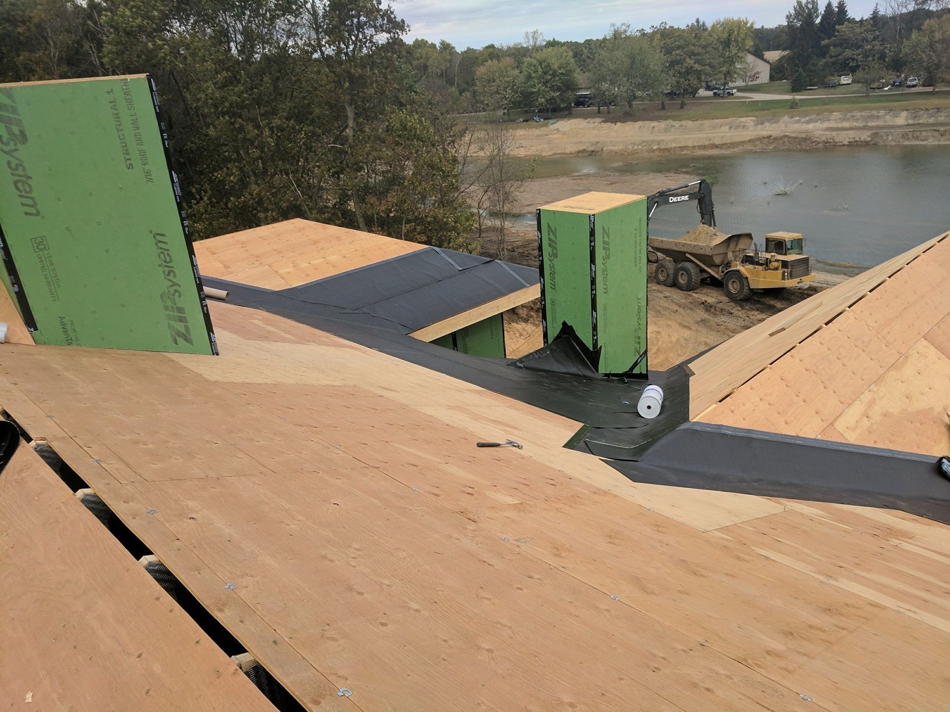 A view from a construction site rooftop showing wooden roof decking, green wall panels, and a yellow excavator by a lake.