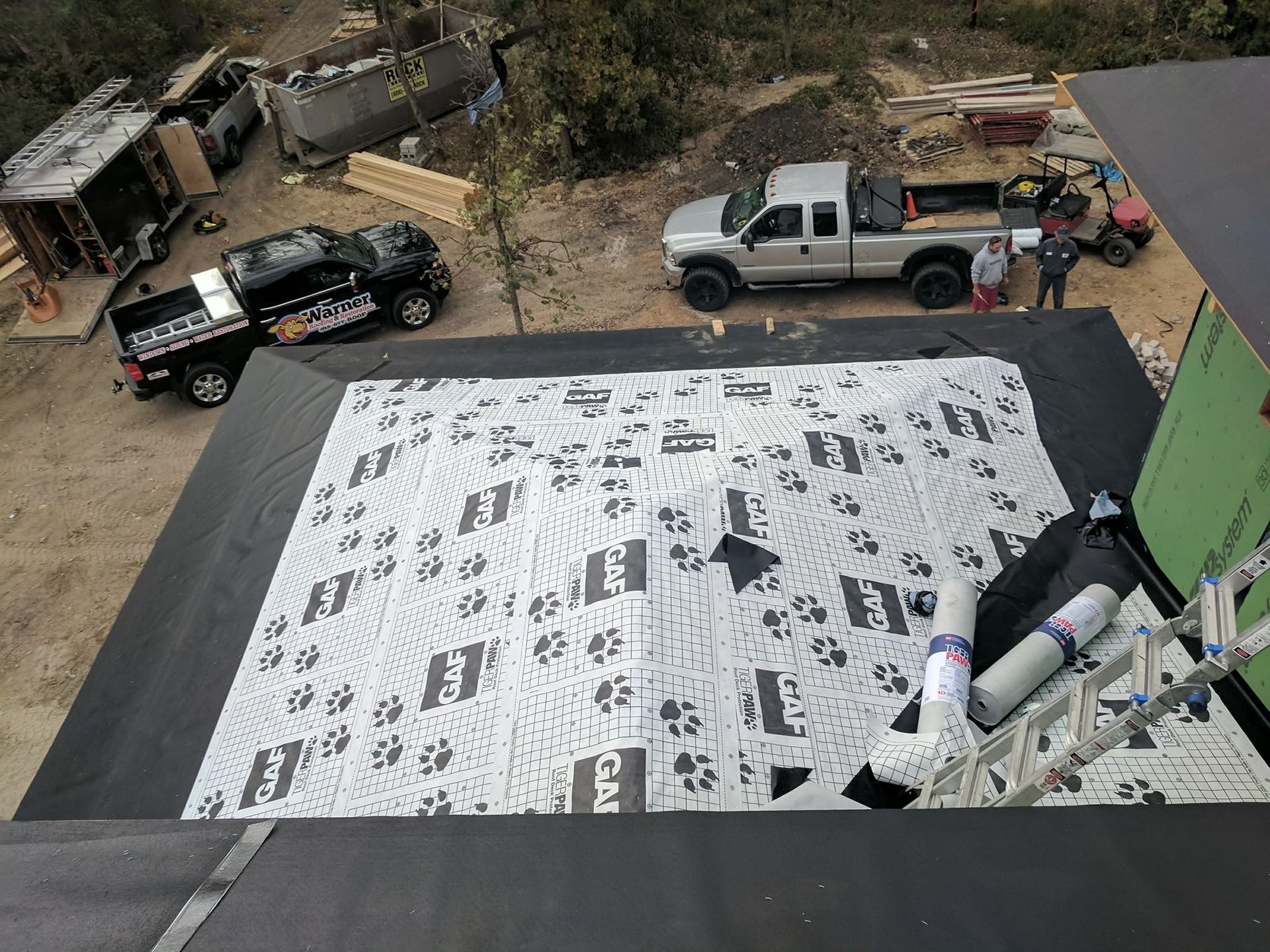 An elevated view of a building roof under construction, covered in patterned underlayment, with vehicles parked nearby.