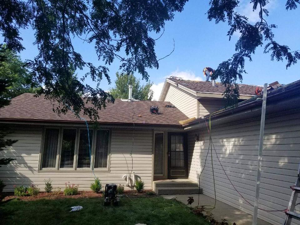 A person works on the roof of a beige house with brown shingles on a sunny day, with a ladder visible in the foreground.