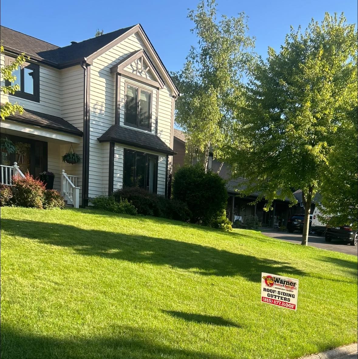 A two-story, cream-colored house with dark trim sits on a sloped green lawn with a real estate sign in the front yard.