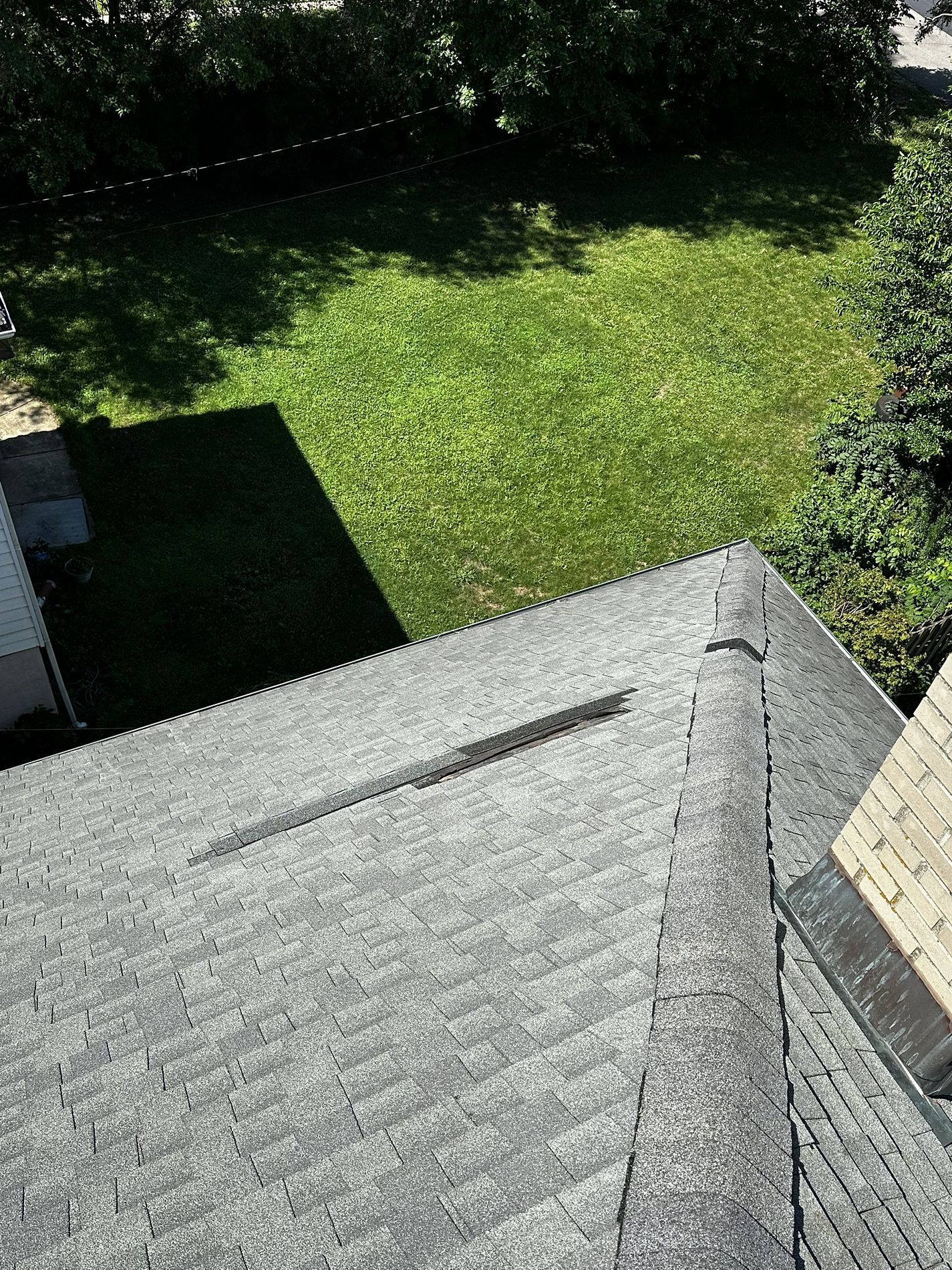 An aerial view of a gray asphalt shingle roof with a ridge vent and a chimney, overlooking a green backyard.