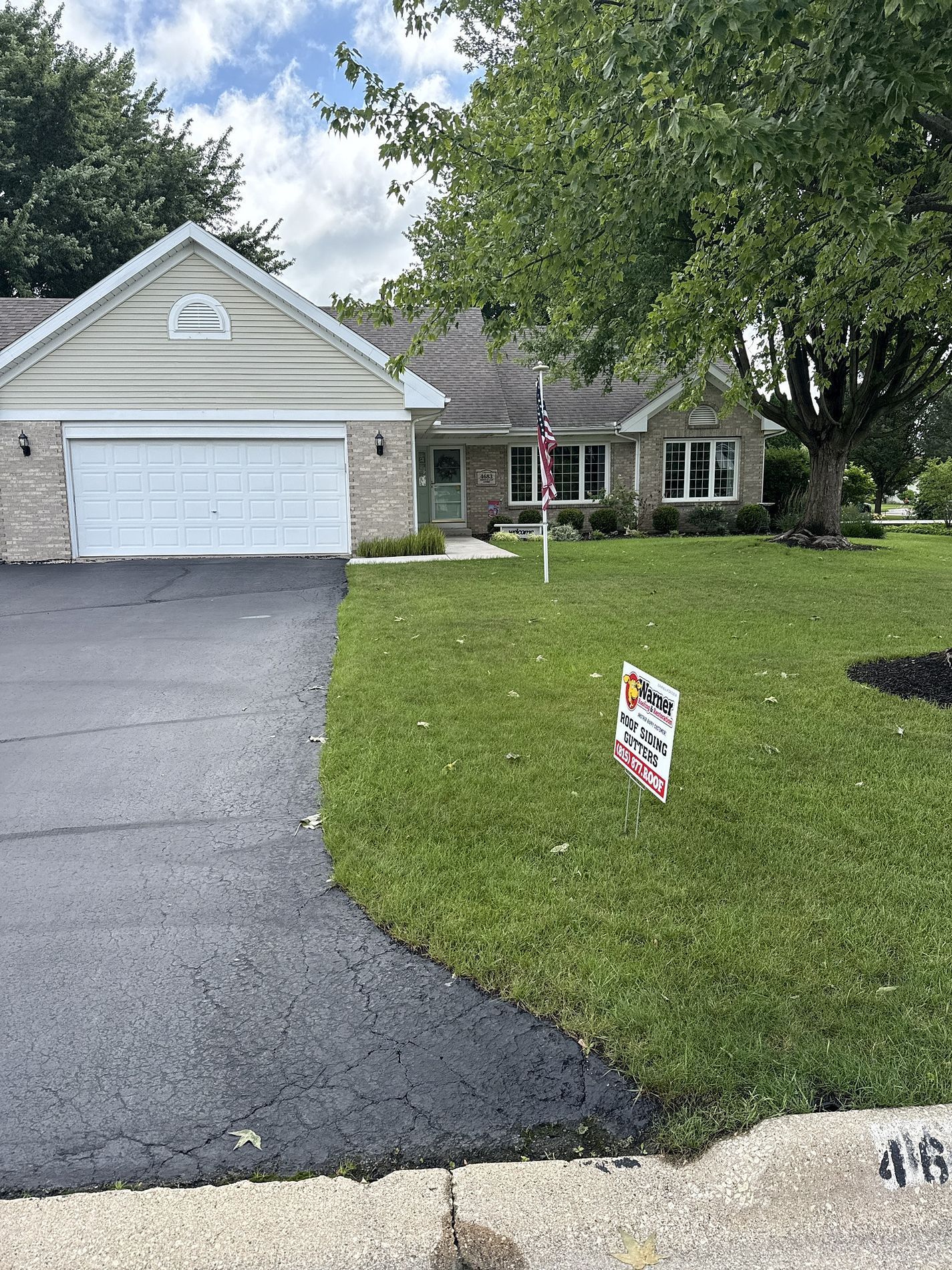 A light-colored house with a single-car garage and asphalt driveway sits behind a green lawn with a yard sign.