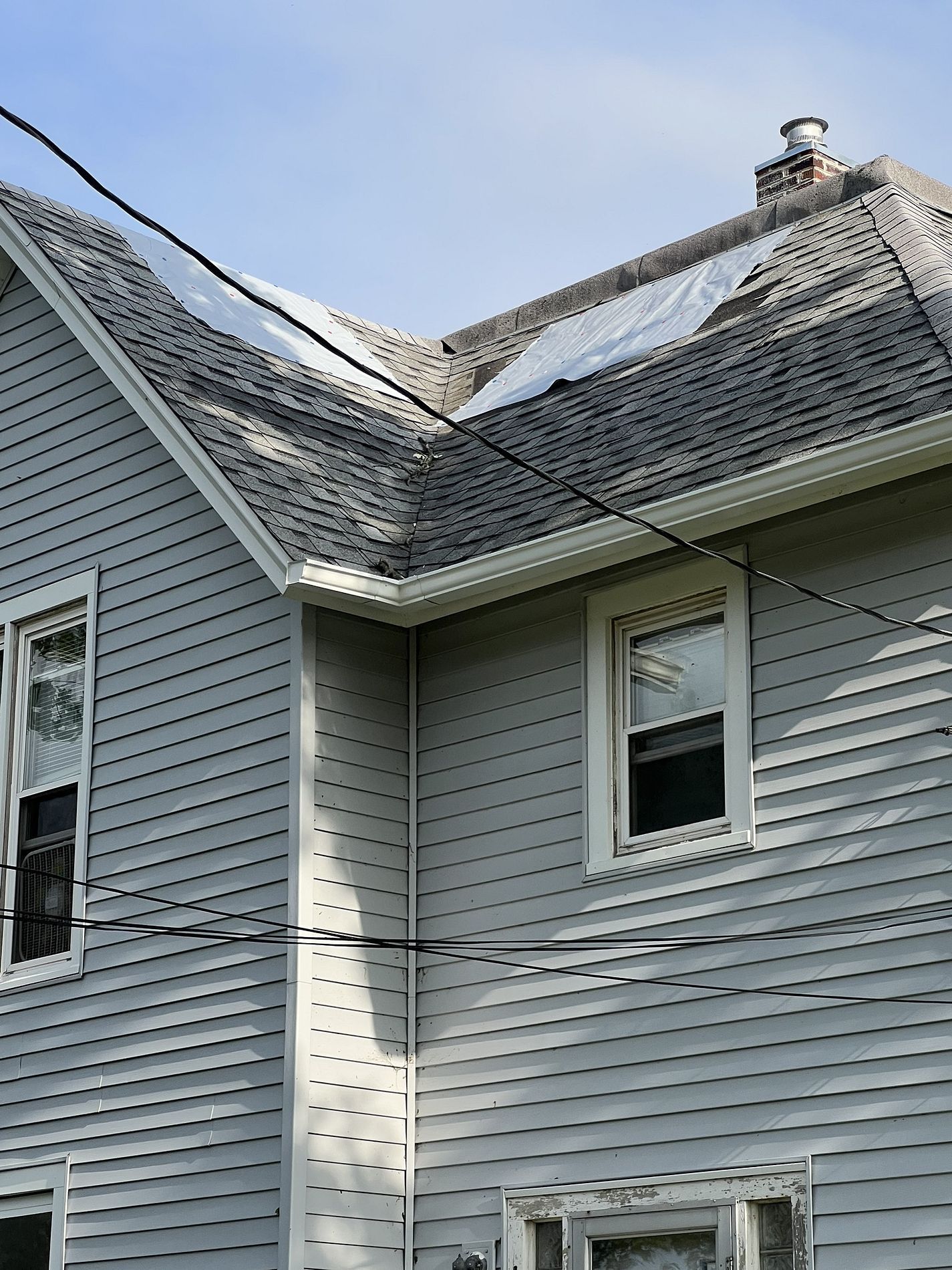 A gray-sided house exterior featuring two roof sections with white tarps covering damaged areas under a blue sky.