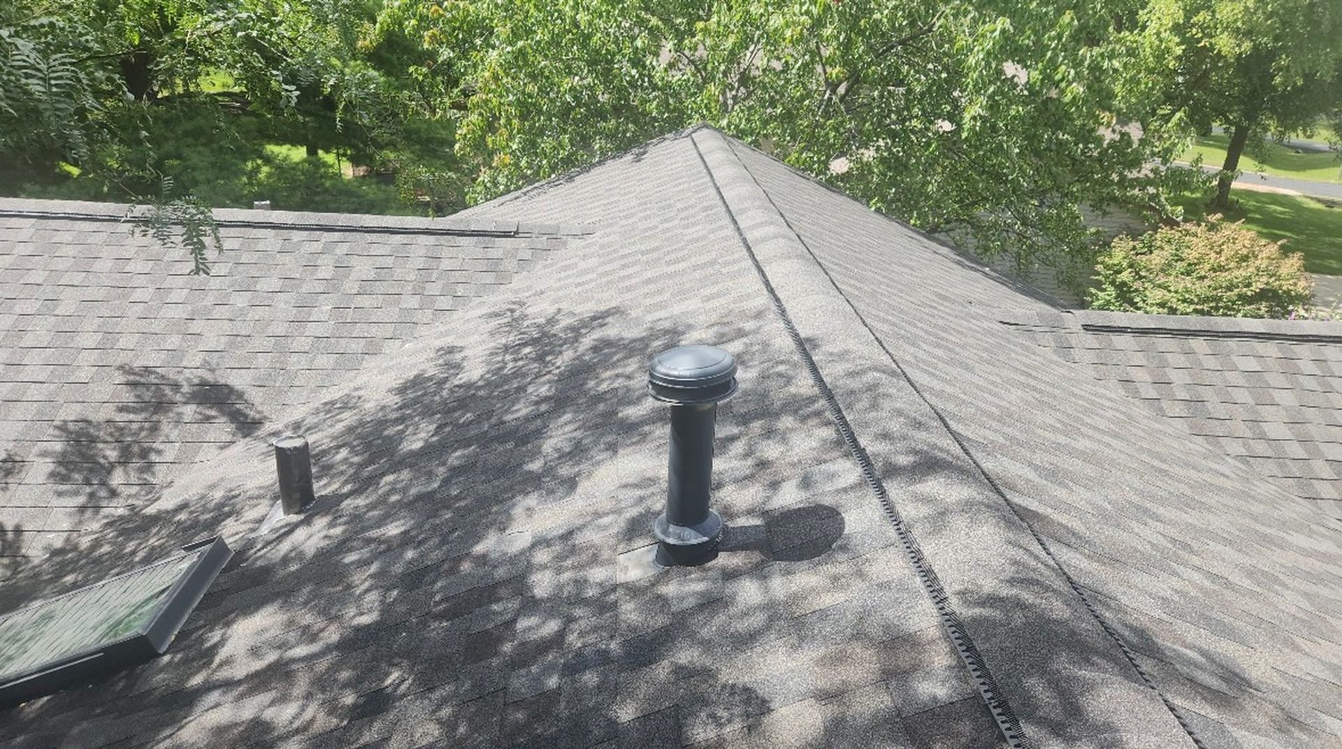 A gray shingled roof with a black plumbing vent pipe and a solar fan unit installed, surrounded by green trees.
