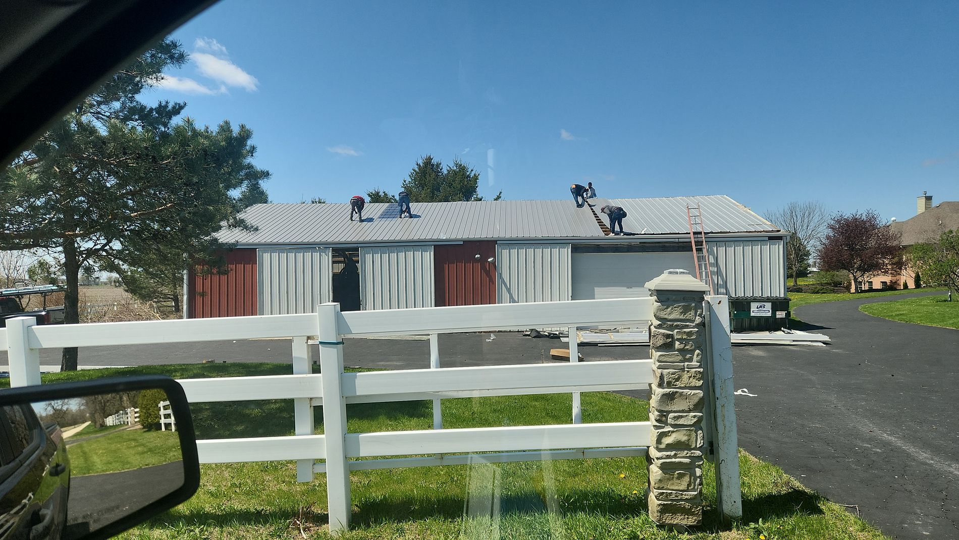 Workers on a ladder and on the roof are installing a metal roof on a long barn with red and white siding.