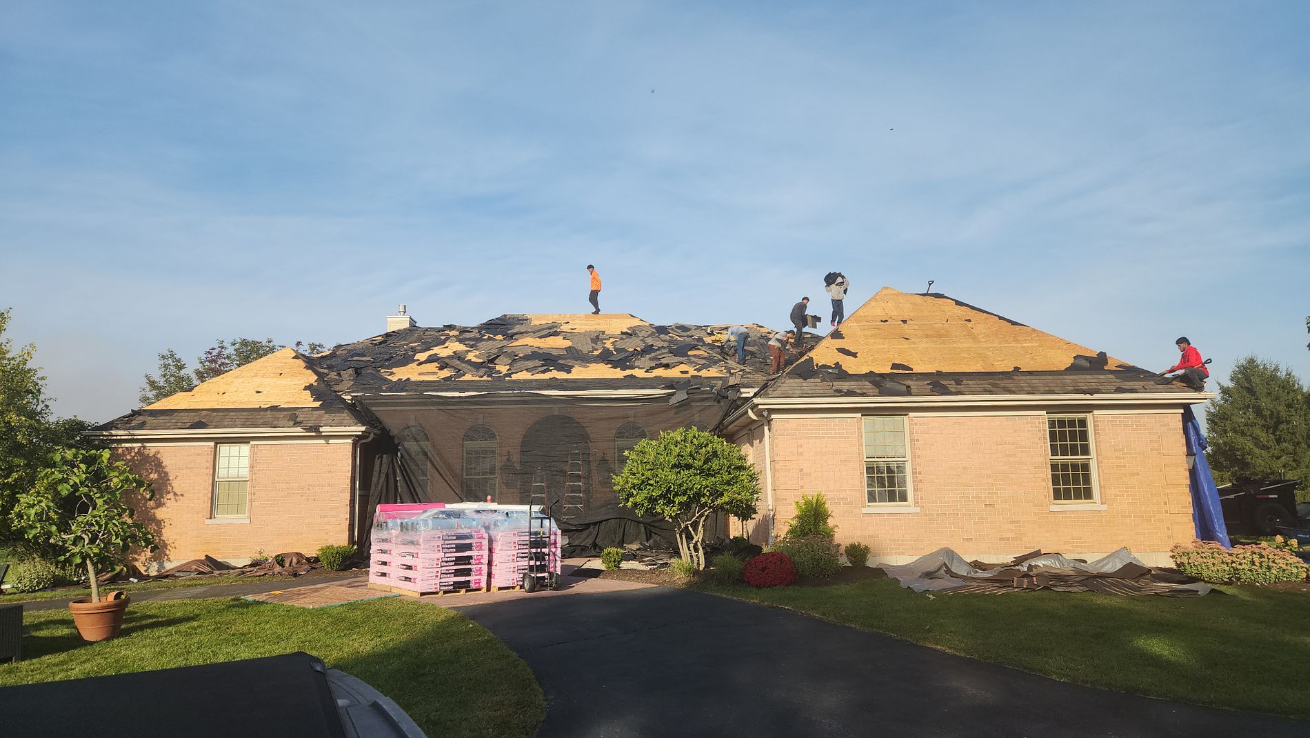 Workers on the roof of a beige brick house replace shingles, with stacks of roofing materials on the driveway.