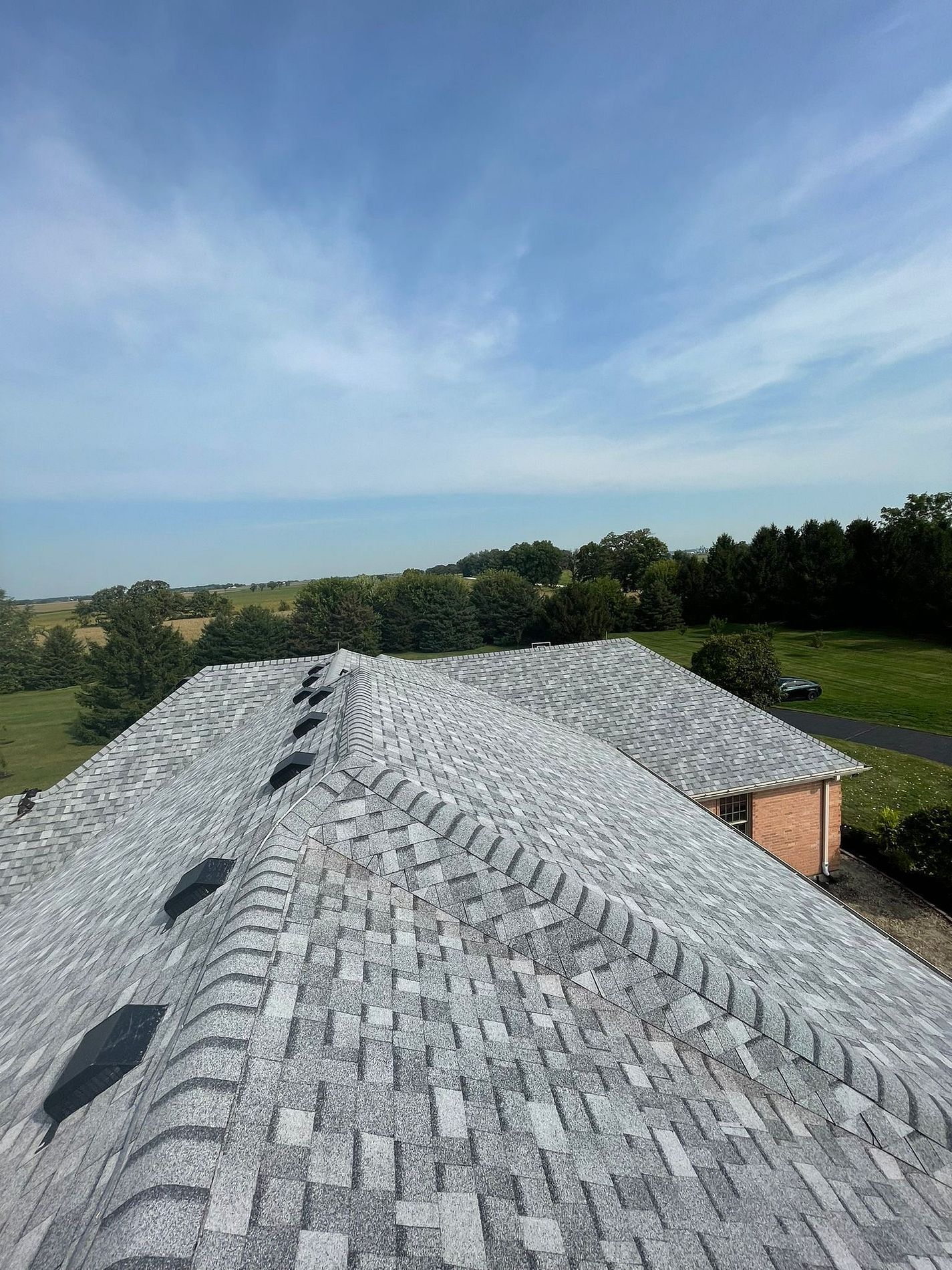 A view from above a multi-gabled gray shingled roof under a blue sky, overlooking a rural landscape with trees and fields.