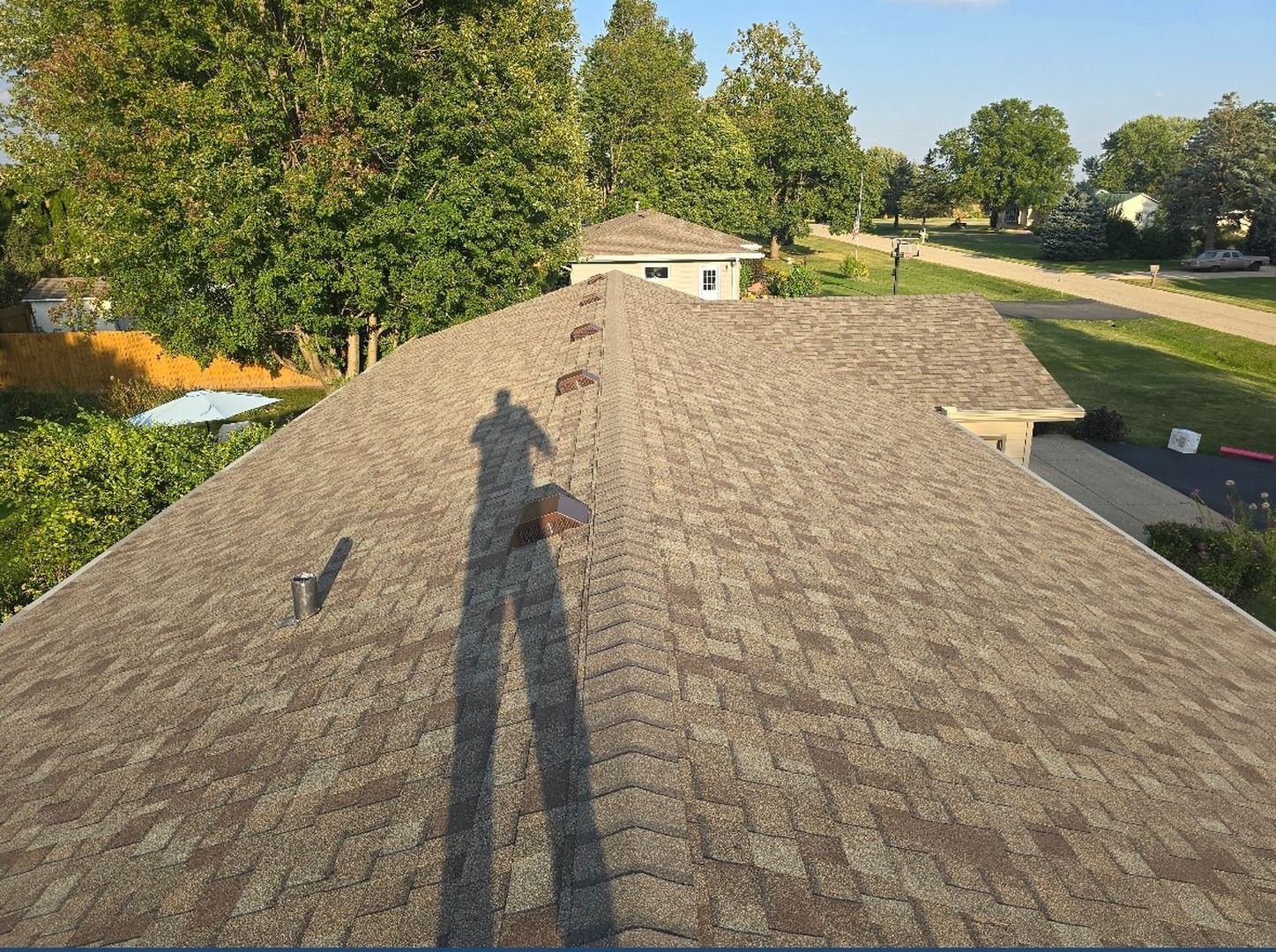 A high-angle view of a brown asphalt shingled roof with a person's shadow cast across the center on a sunny day.