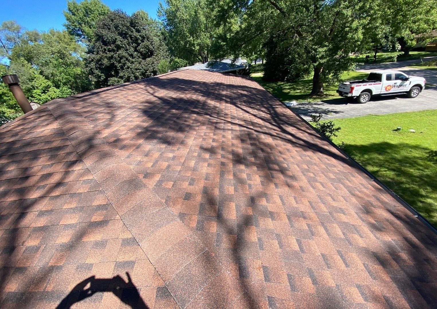 An angled view of a textured brown asphalt shingle roof, with trees and a white pickup truck in the background.