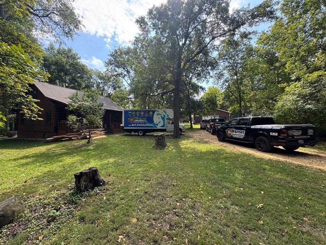 A moving truck and a pickup truck parked on a grass-covered driveway beside a house surrounded by trees.