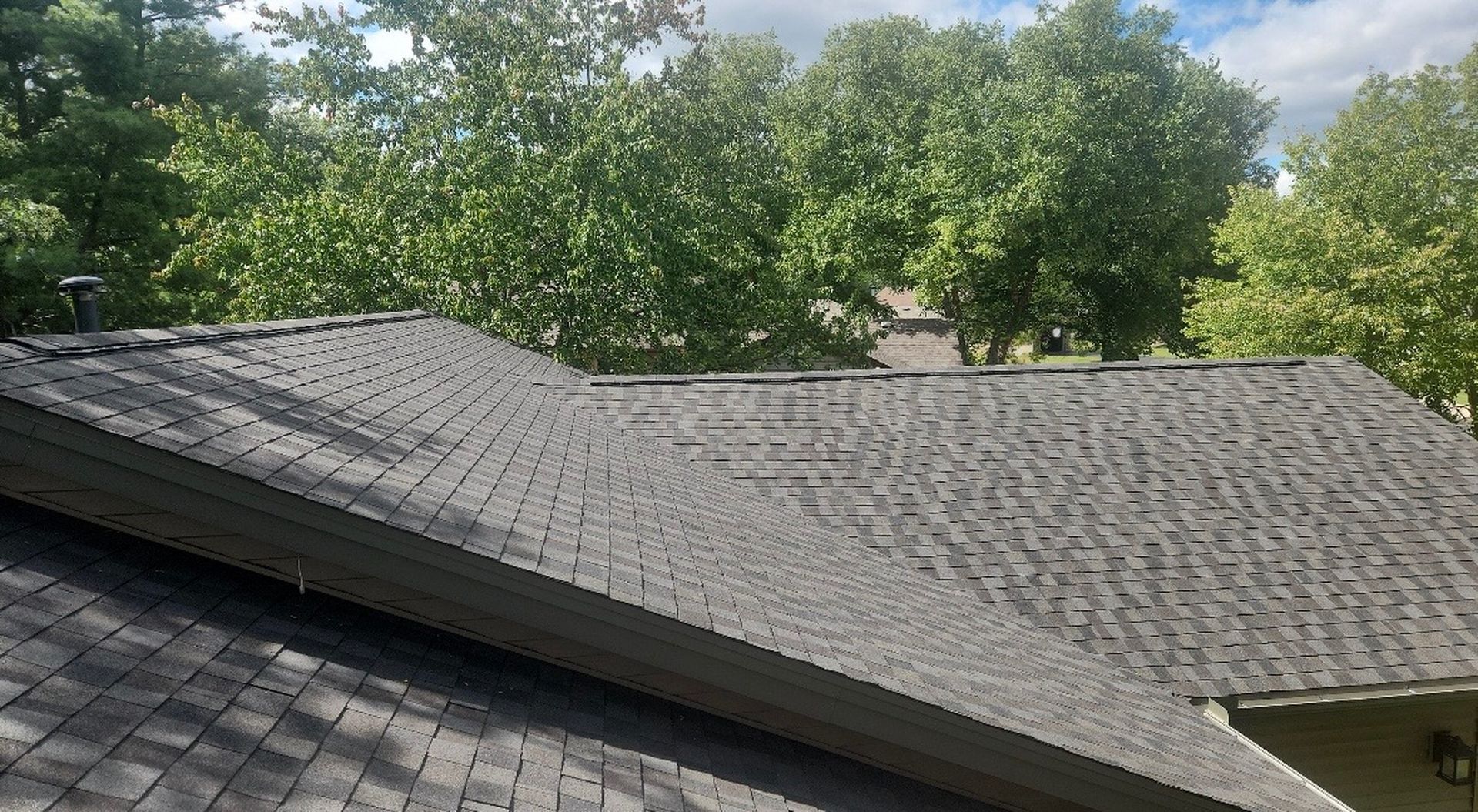 A view of a shingled residential roof with a valley and ridge vents, framed by green trees under a partly cloudy sky.