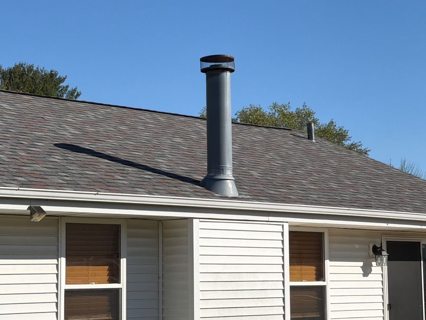 A gray metal chimney stack and a smaller pipe extending from the shingled roof of a house with light-colored siding.