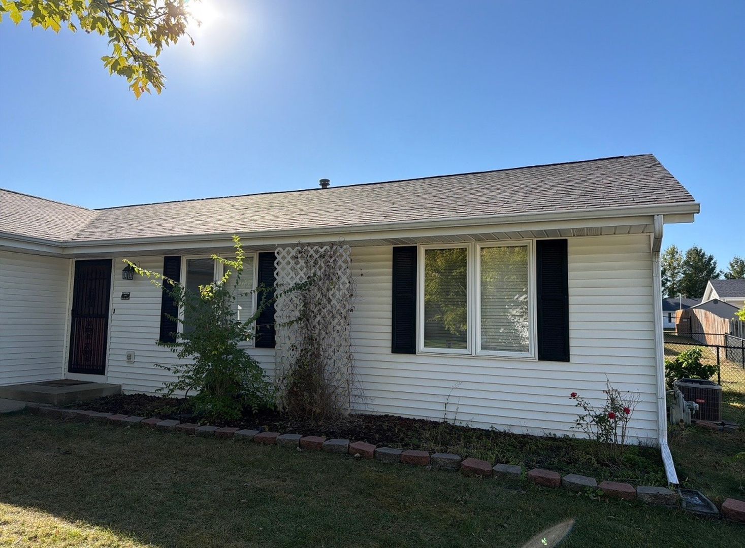 A single-story ranch house with white horizontal siding, black window shutters, and a shingled roof under a clear blue sky.
