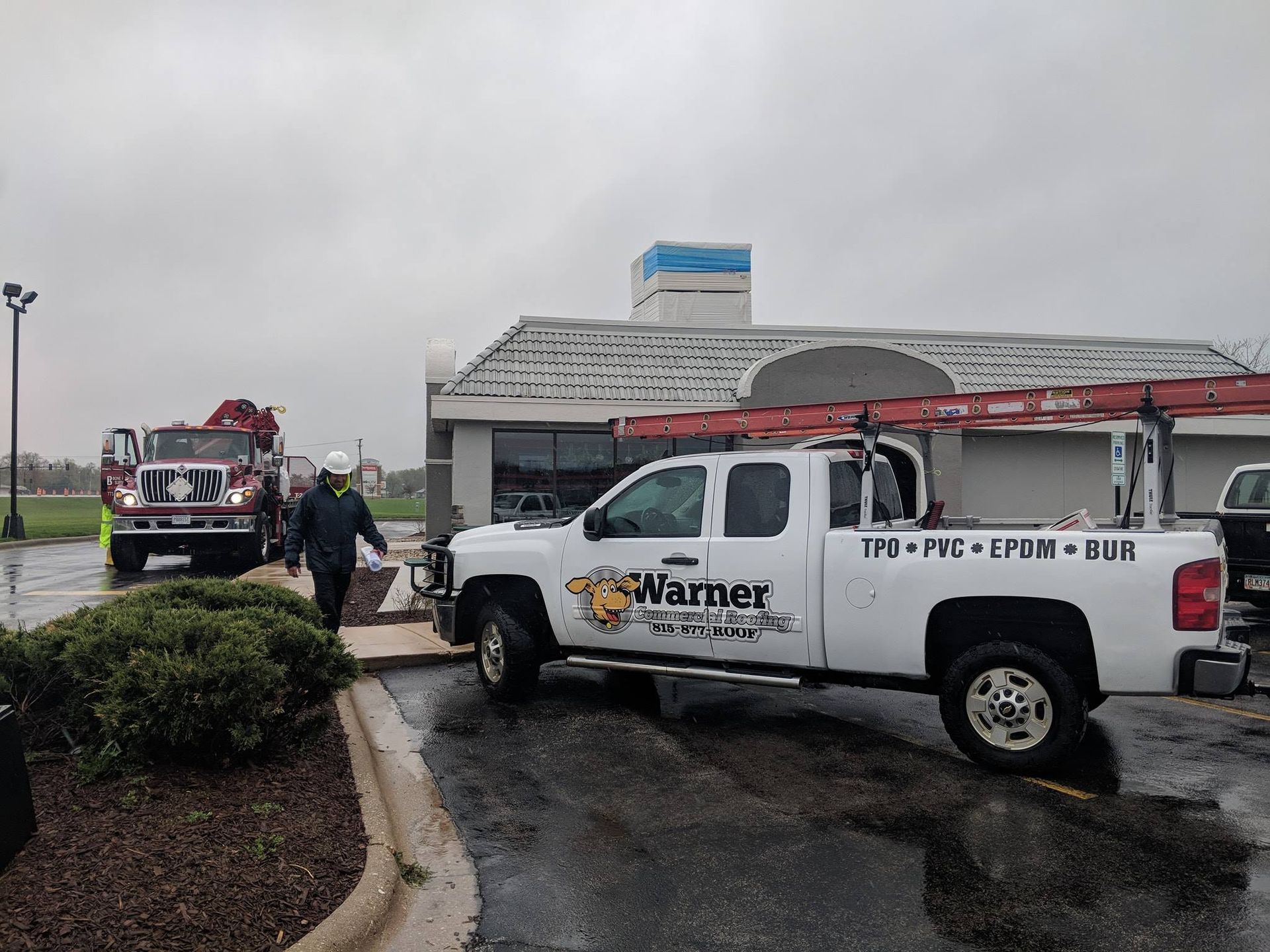 A white Warner Roofing truck and a fire truck parked at a building site during the day.