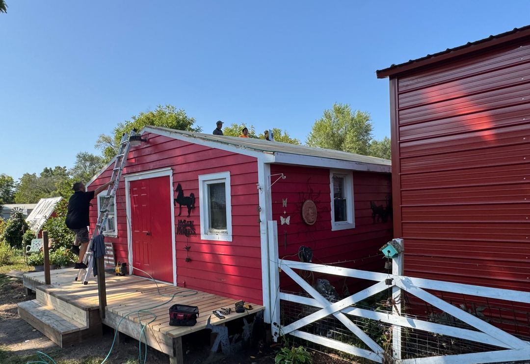 A person works on a ladder near the wooden porch of a small red shed with white trim on a sunny day.