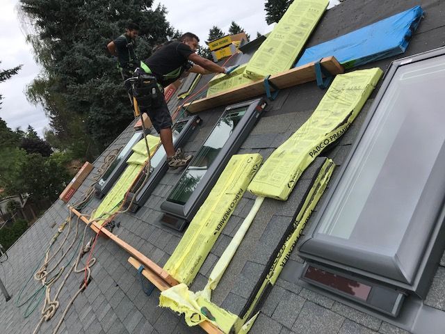Two workers on a residential roof installing skylights, using wooden planks for stability and ropes for safety.