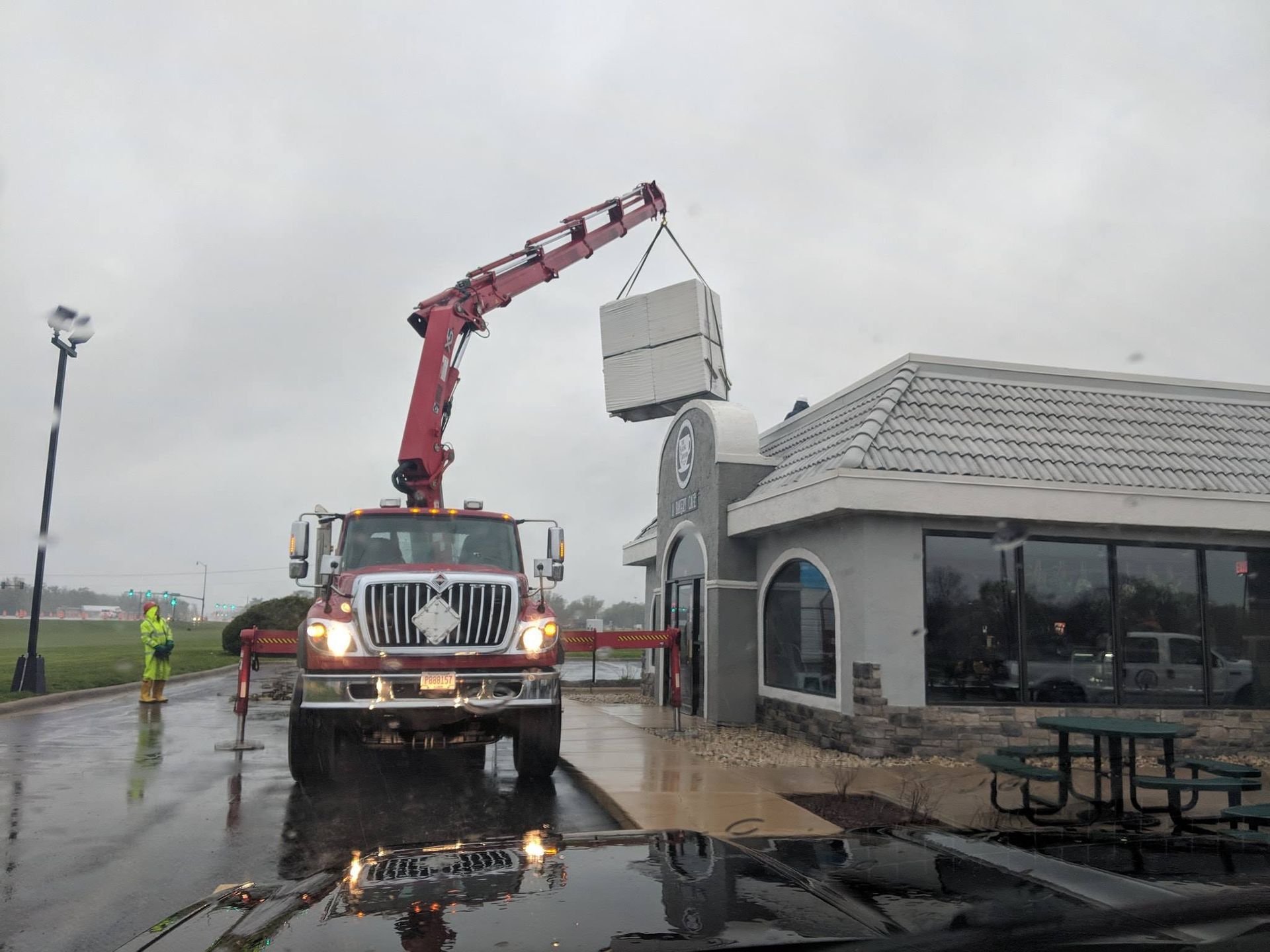 A red crane truck lifts a large HVAC unit onto the roof of a commercial building on a rainy day.