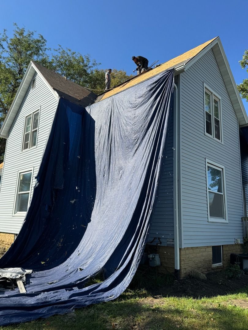 A worker stands on the roof of a blue siding house, with a large blue tarp covering a section of the roof and side wall.