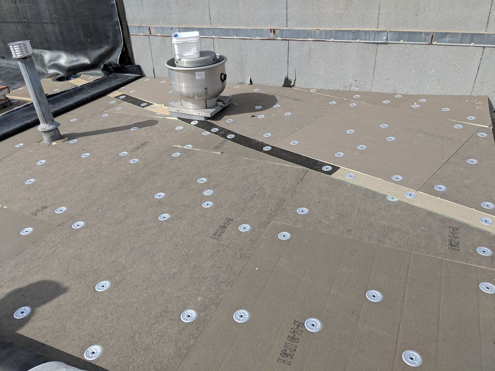 A flat commercial roof showing installed insulation boards with numerous circular fasteners and a metal ventilation unit.