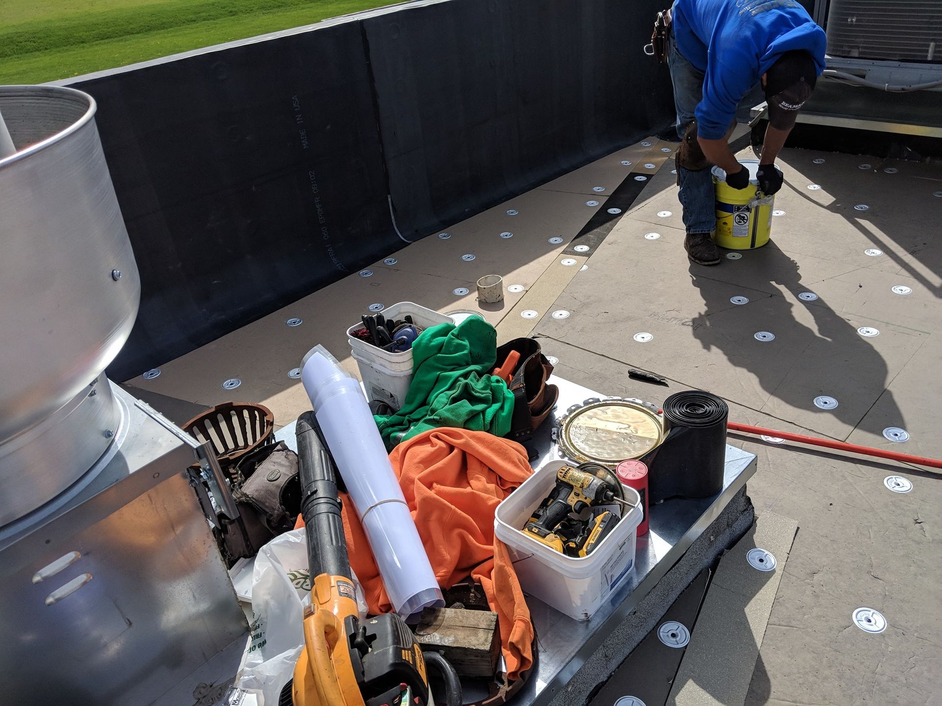 A worker in a blue shirt on a rooftop holds a yellow container near a collection of construction tools and supplies.