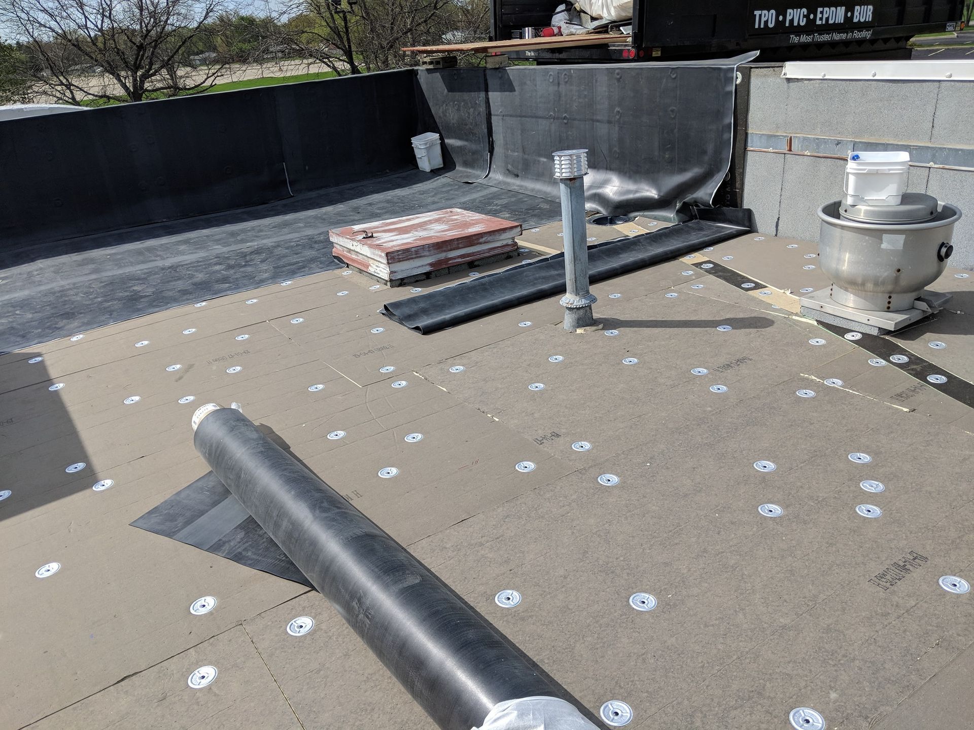 A rooftop under construction with insulation boards, roofing material rolls, a metal vent, and a dumpster in the background.